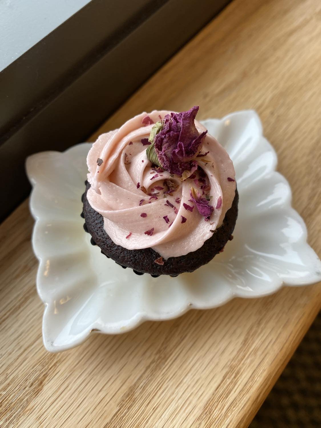 Chocolate cupcake with pink frosting topped with dried rose petals, placed on a decorative white plate on a wooden surface.