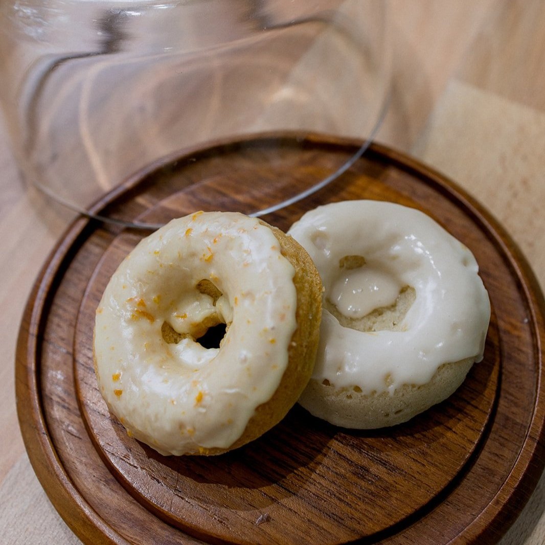Two glazed donuts on a wooden serving board with a glass cover in the background.
