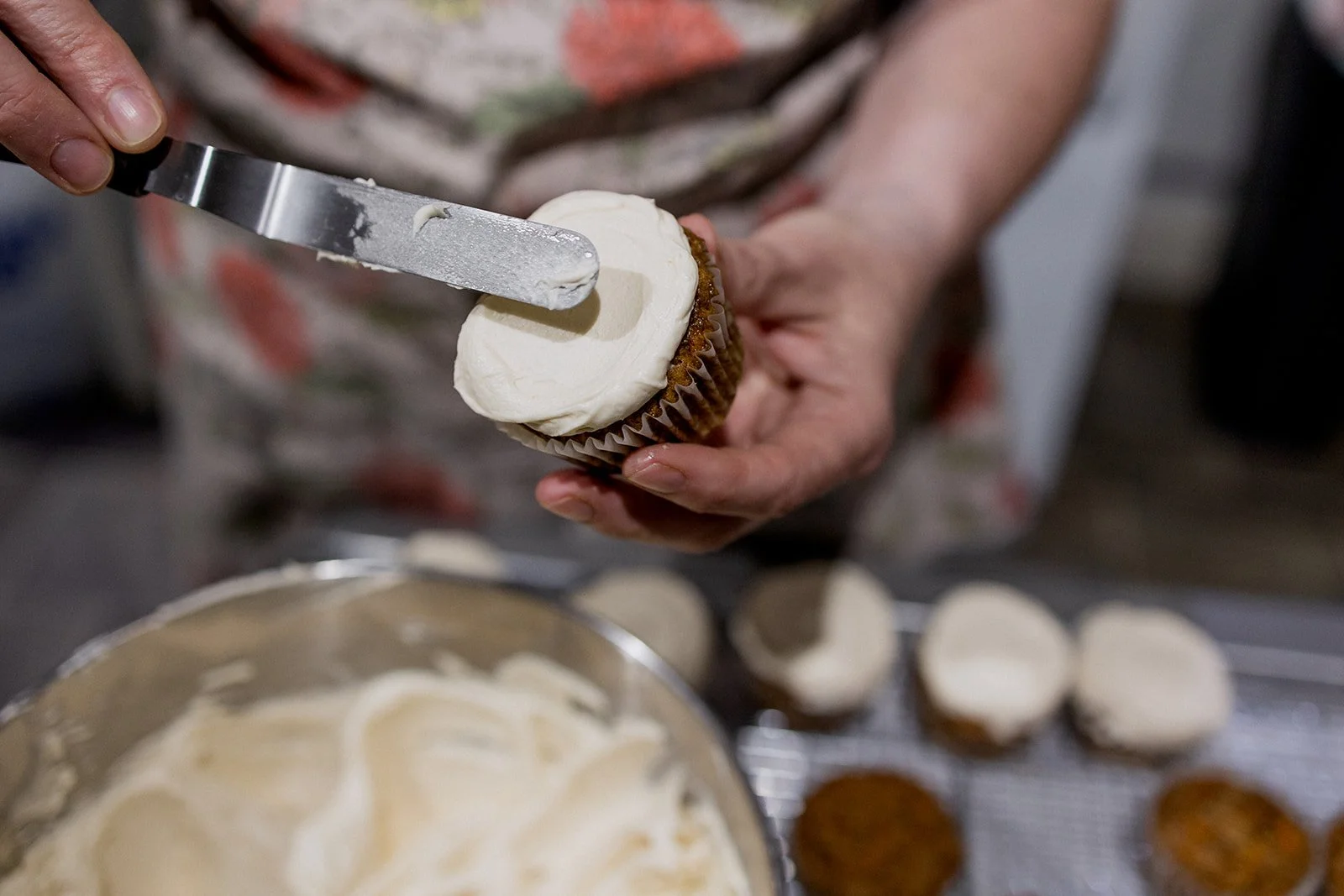 Person decorating a cupcake with white frosting using a spatula, with other cupcakes on a cooling rack in the background.