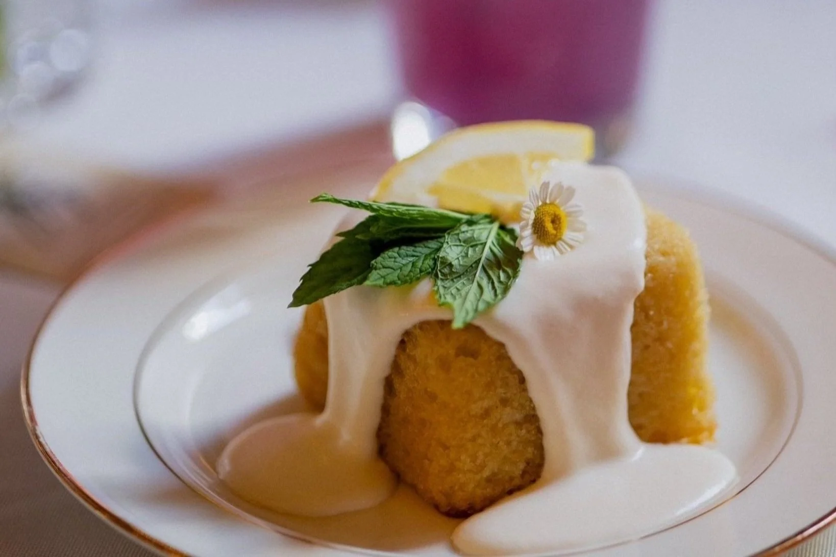 A dessert with a cornbread pudding topped with white sauce, garnished with mint leaves, a lemon wedge, and a small daisy flower on a white plate.