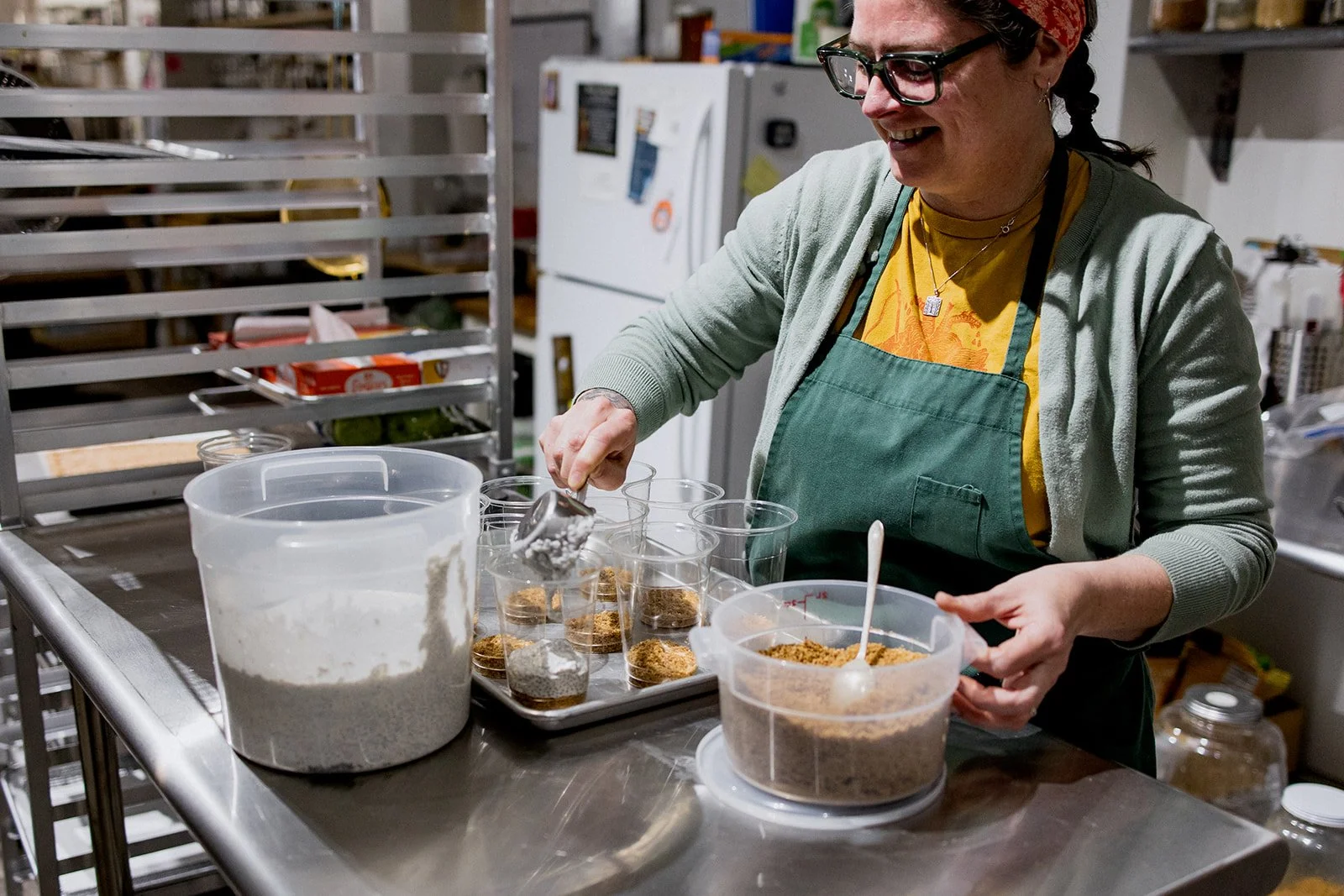 A woman in glasses and a green apron is filling small clear cups with brown and gray powdered ingredients in a kitchen.
