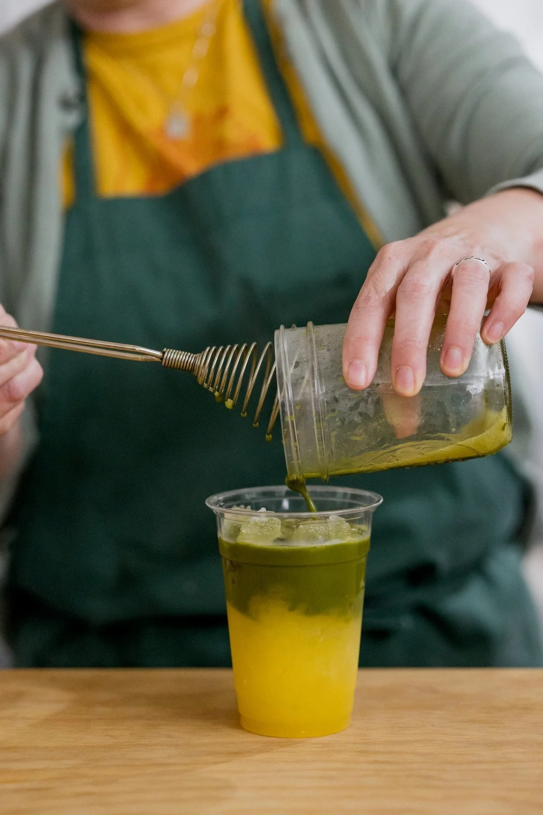 Person pouring matcha green tea into a plastic cup with ice on a wooden table.