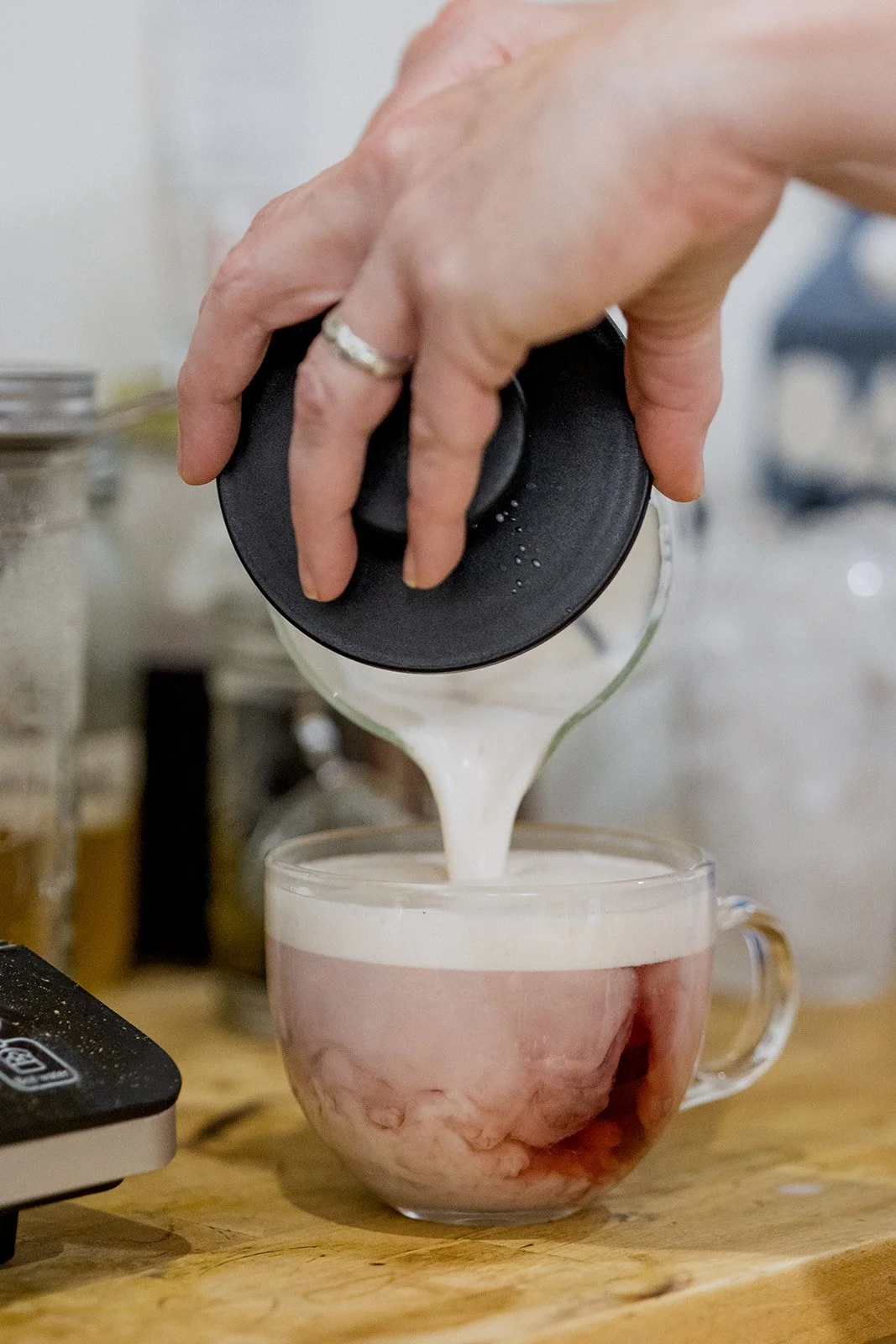 A person pouring a creamy pink beverage from a glass container into a clear glass mug with a handle, on a wooden surface.