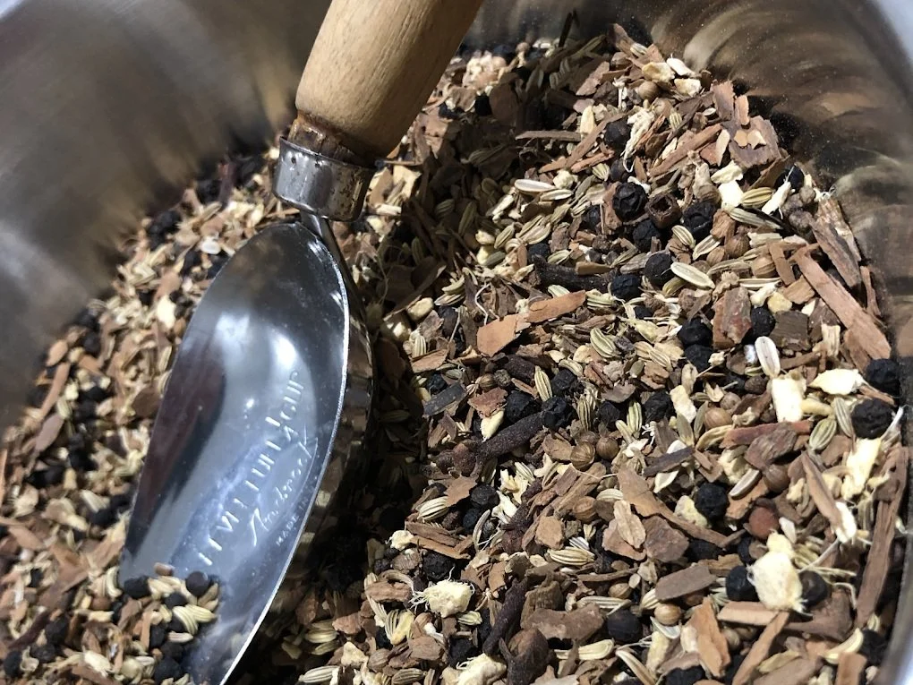 A close-up of a metal container filled with dried herbs, spices, and seeds, with a metal scoop partially inserted.