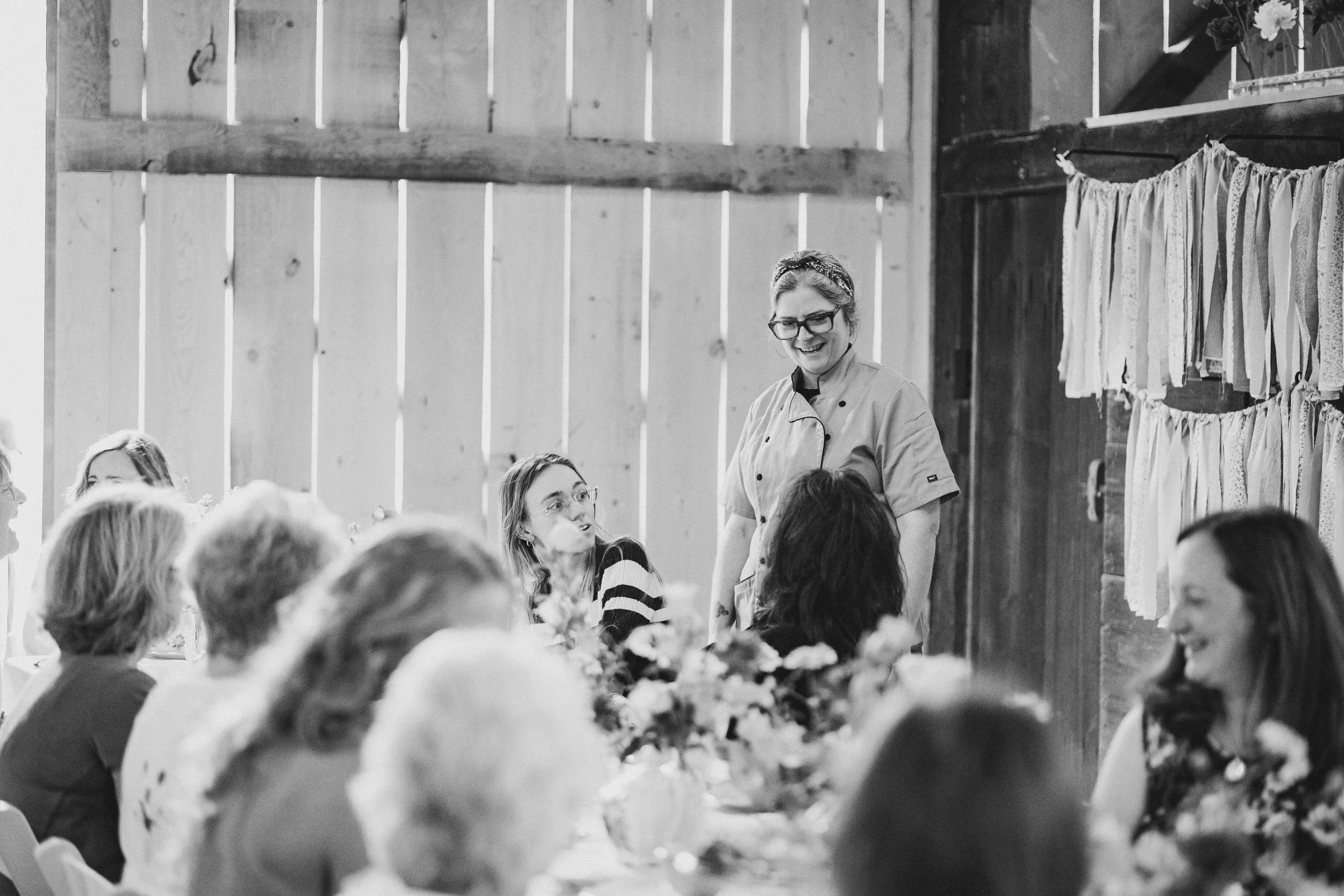 A woman chef speaking to a group of women at a gathering in a rustic setting, with wooden walls and hanging fabric in the background.