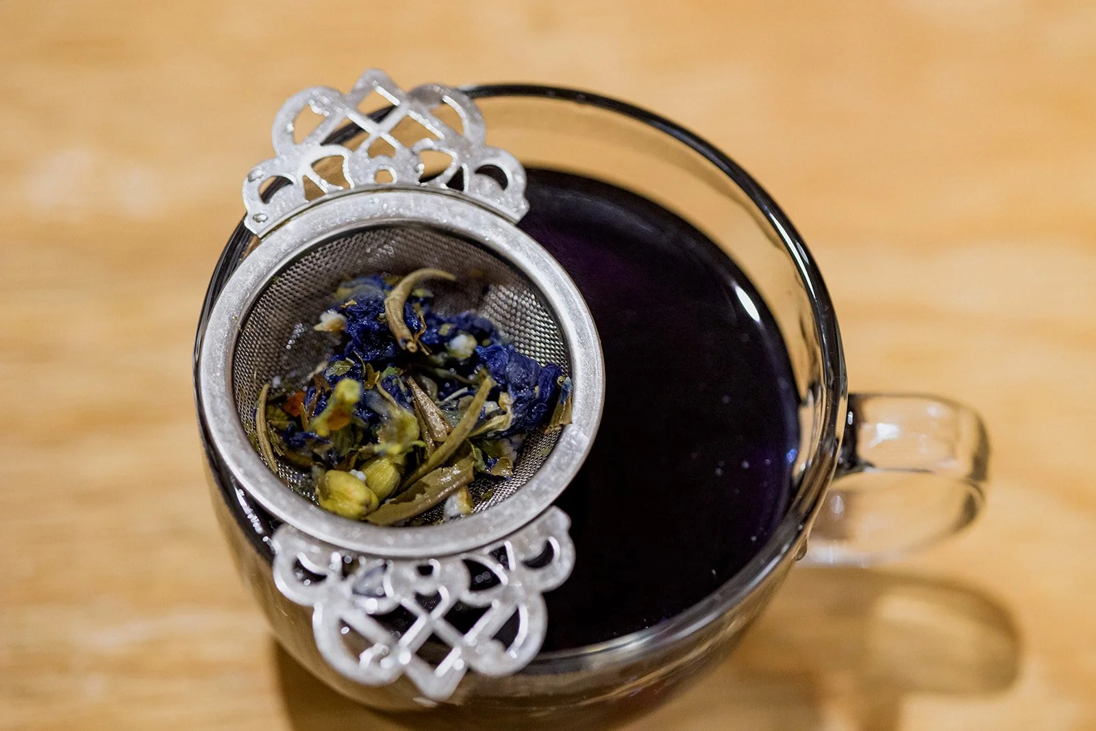 A glass cup filled with dark purple tea, with a metal infuser containing dried blue and green flowers and herbs placed on top of the cup.