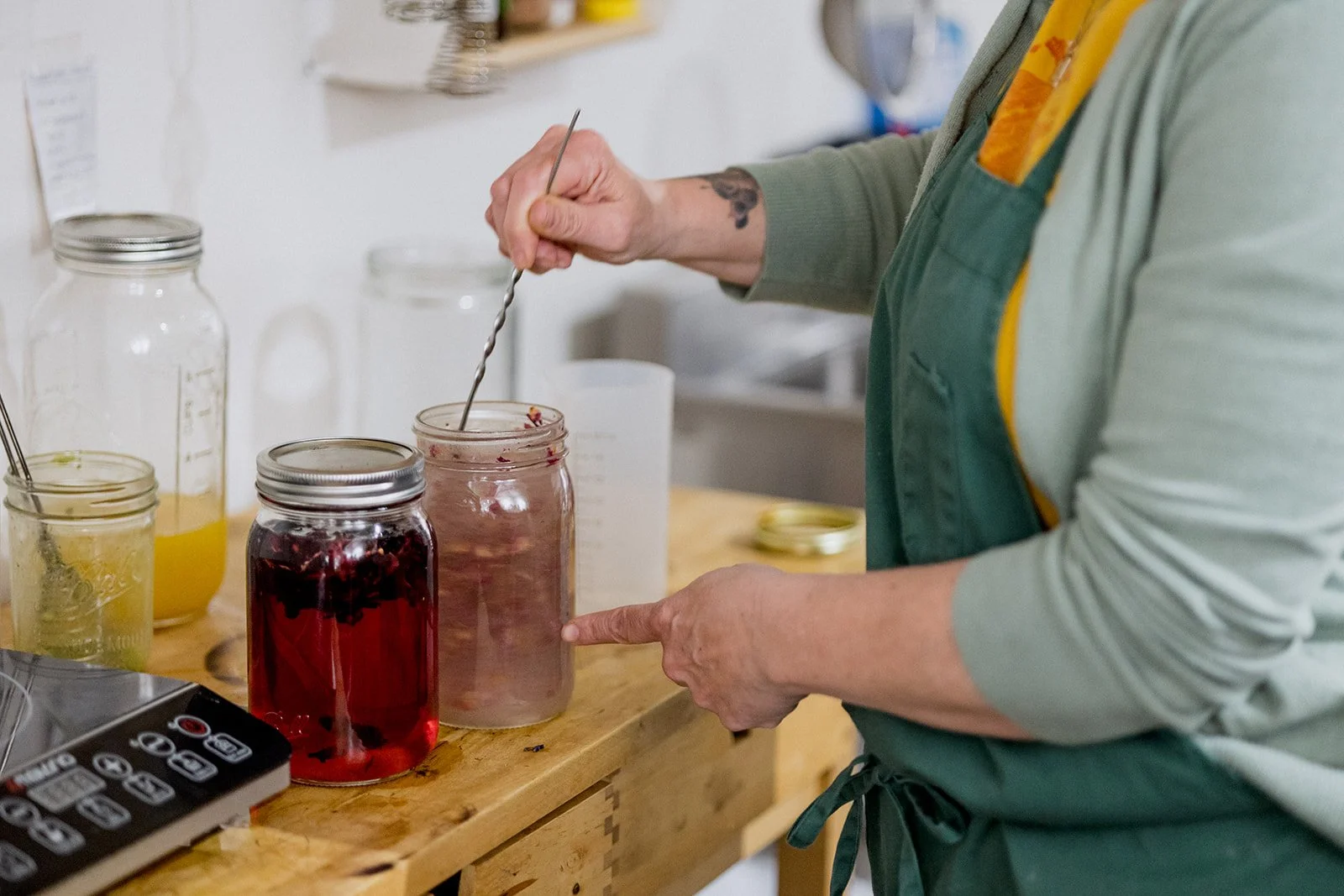 Person stirring a jar filled with a pinkish liquid with a metal spoon, on a wooden table with jars containing different liquids.