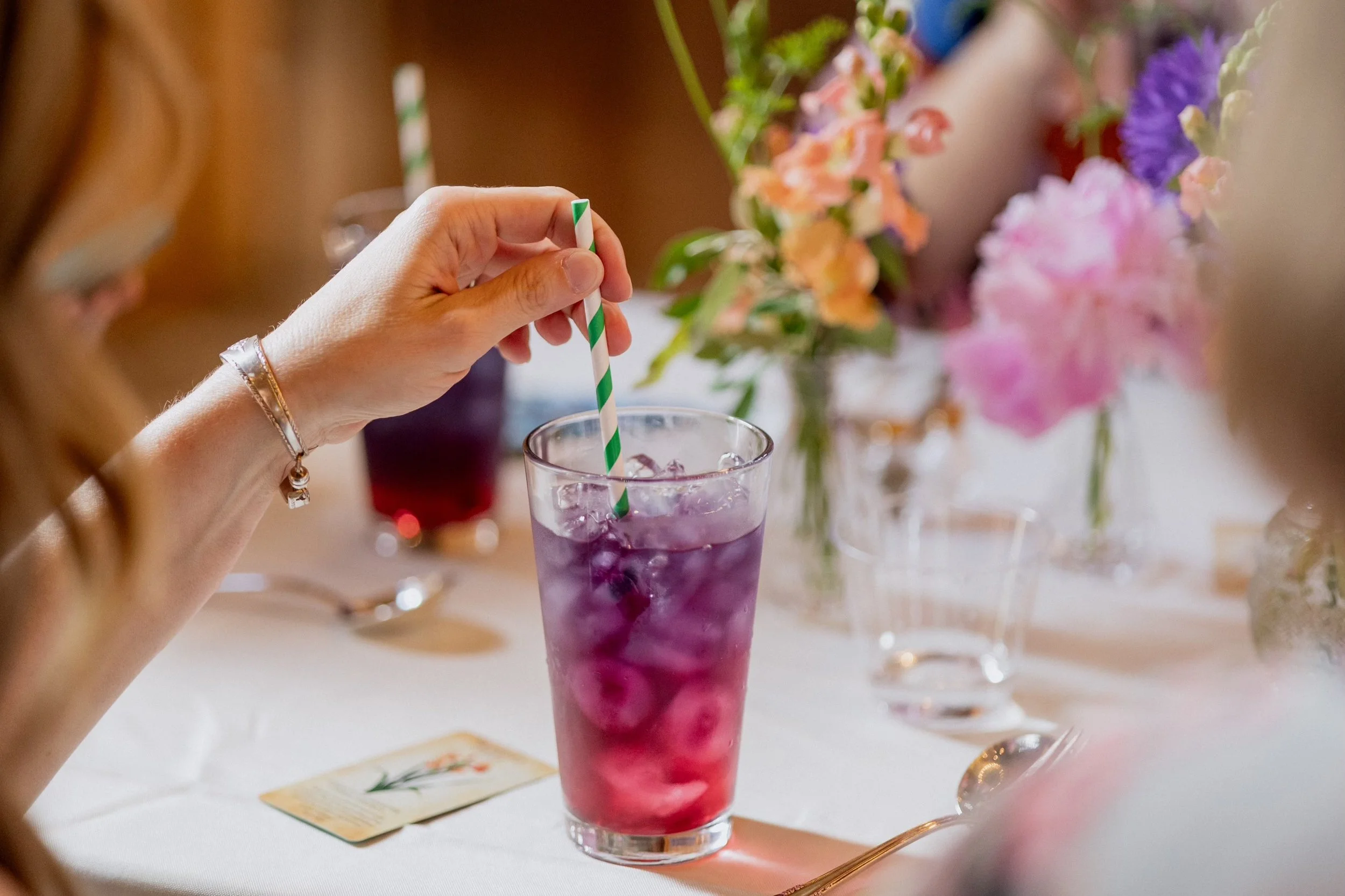 Person stirring a purple drink with ice in a tall glass, on a white table with silverware and floral decorations in the background.