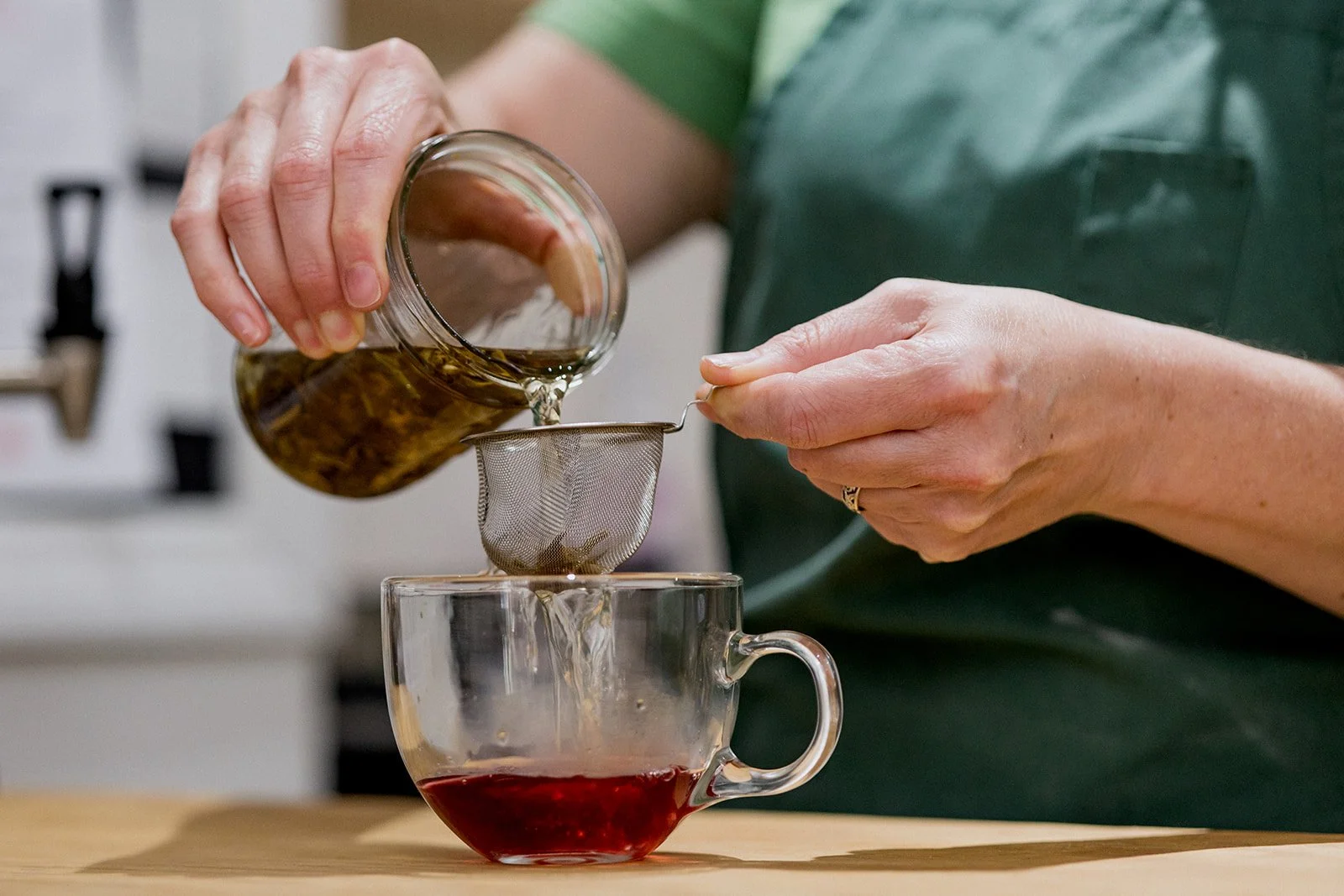 A person wearing a green apron is pouring hot tea from a glass jar through a strainer into a glass cup.