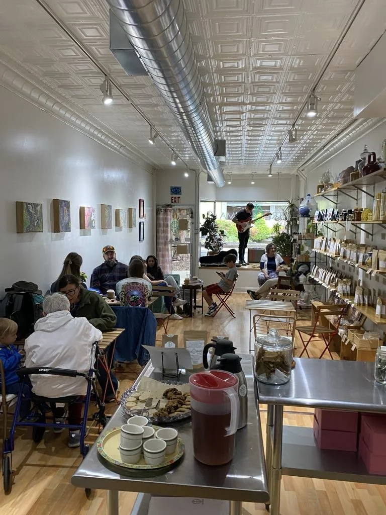 Interior of a cozy cafe with people sitting at tables, a musician playing guitar near the window, shelves with jars and packaged products on the right, and a counter with coffee cups and a pitcher in the foreground.