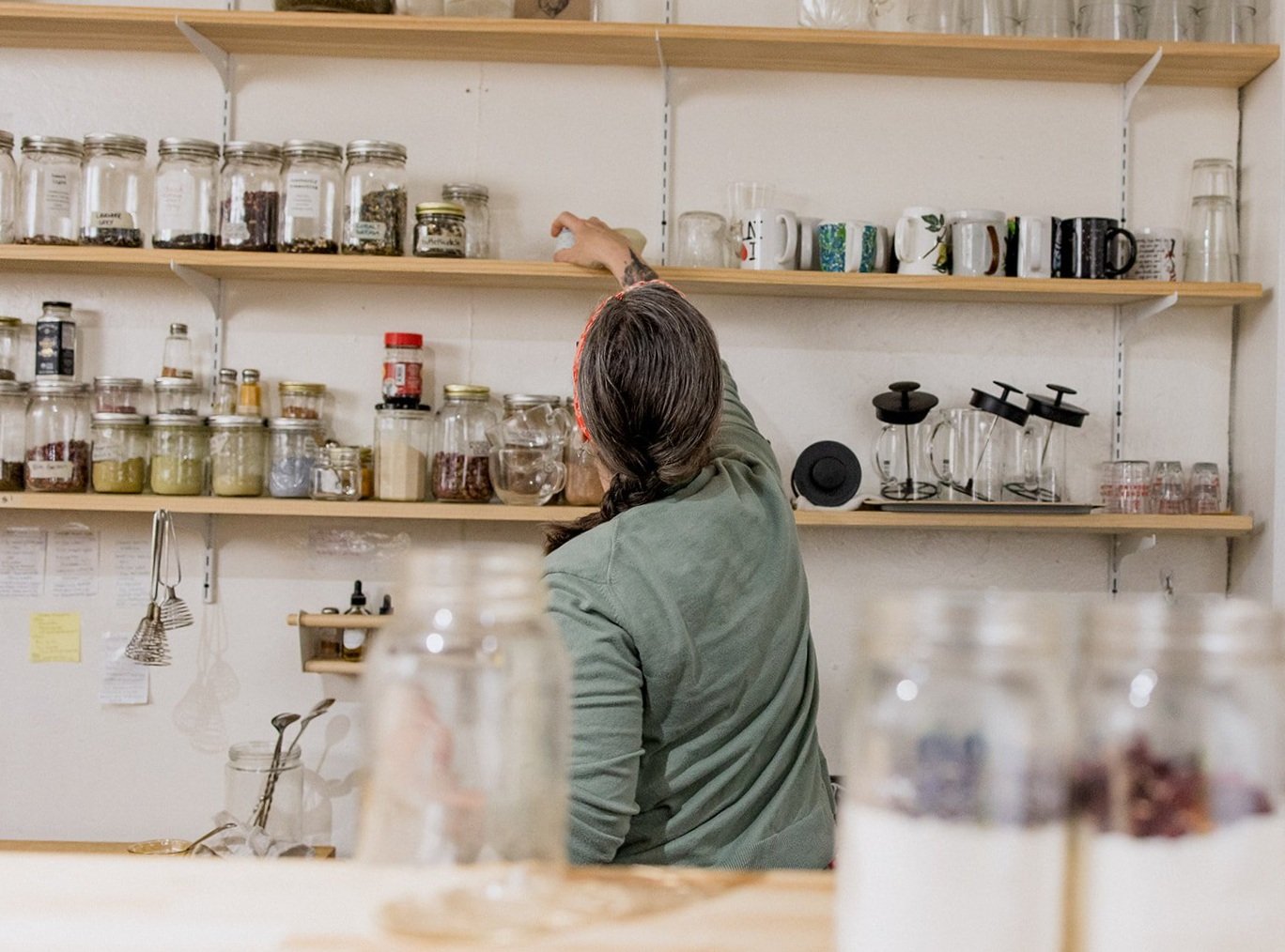 A person with dark hair and a green shirt reaching up to a shelf filled with jars and mugs in a kitchen or coffee shop setting.