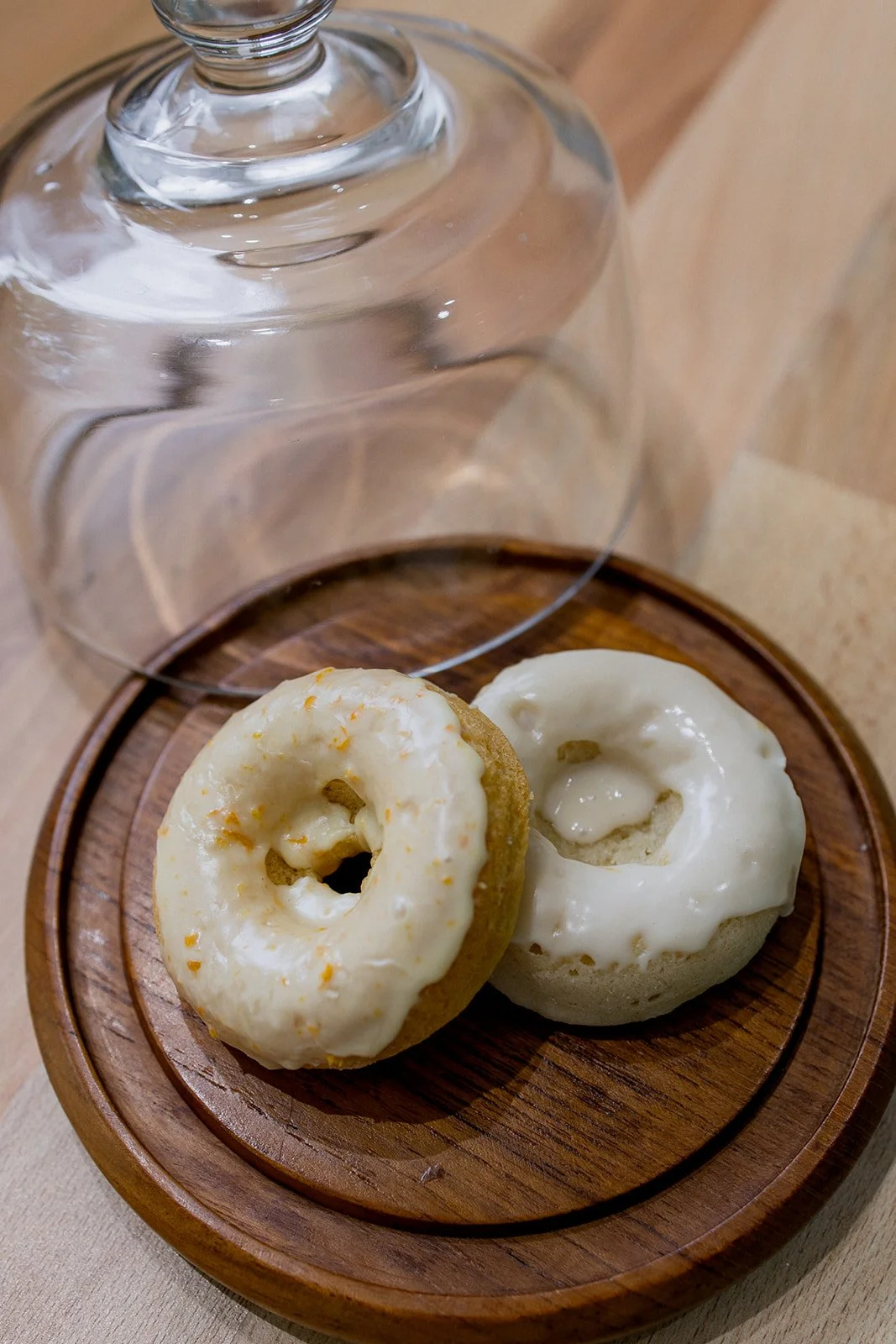 Two donuts with white glaze on a round wooden tray, with a glass cloche cover placed upside down behind them.