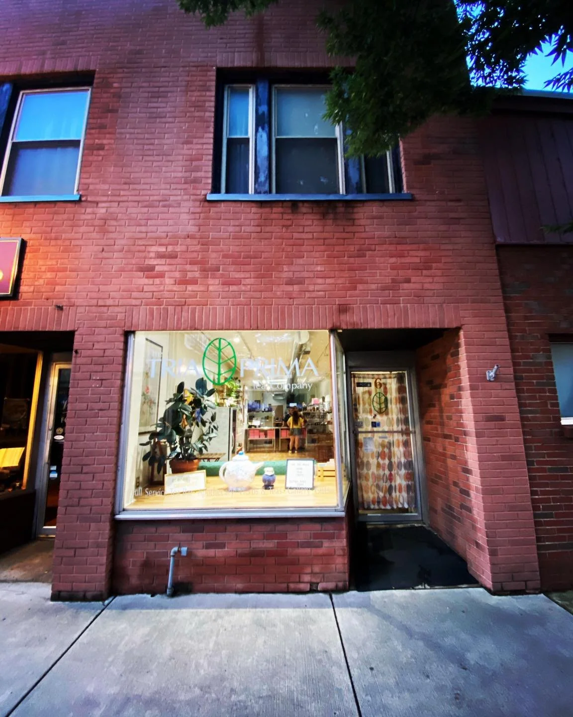 Red brick building with a large front window displaying a plant, teapots, and a sign for Tea Company. Above are two second-floor windows, and the building has a small sign on the left side.
