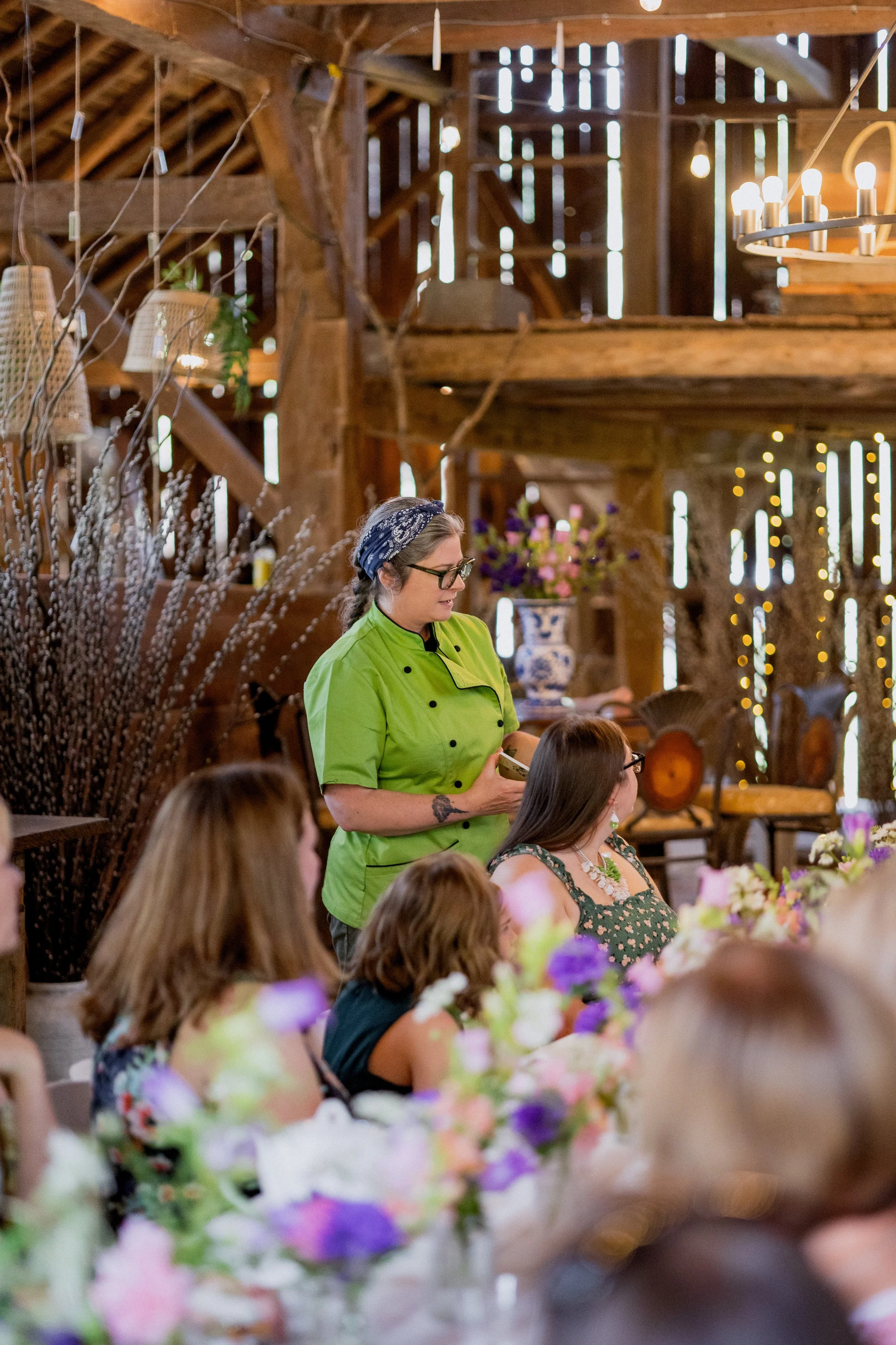 A woman in a green chef's coat and a headscarf stands at a table with seated guests in a rustic barn setting, holding a menu or note, with floral decorations and wooden beams in the background.