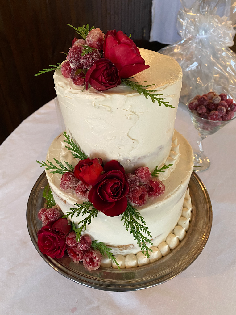 Two-tiered white frosted wedding cake decorated with red roses, ice-covered red berries, and green foliage, placed on a silver cake stand.
