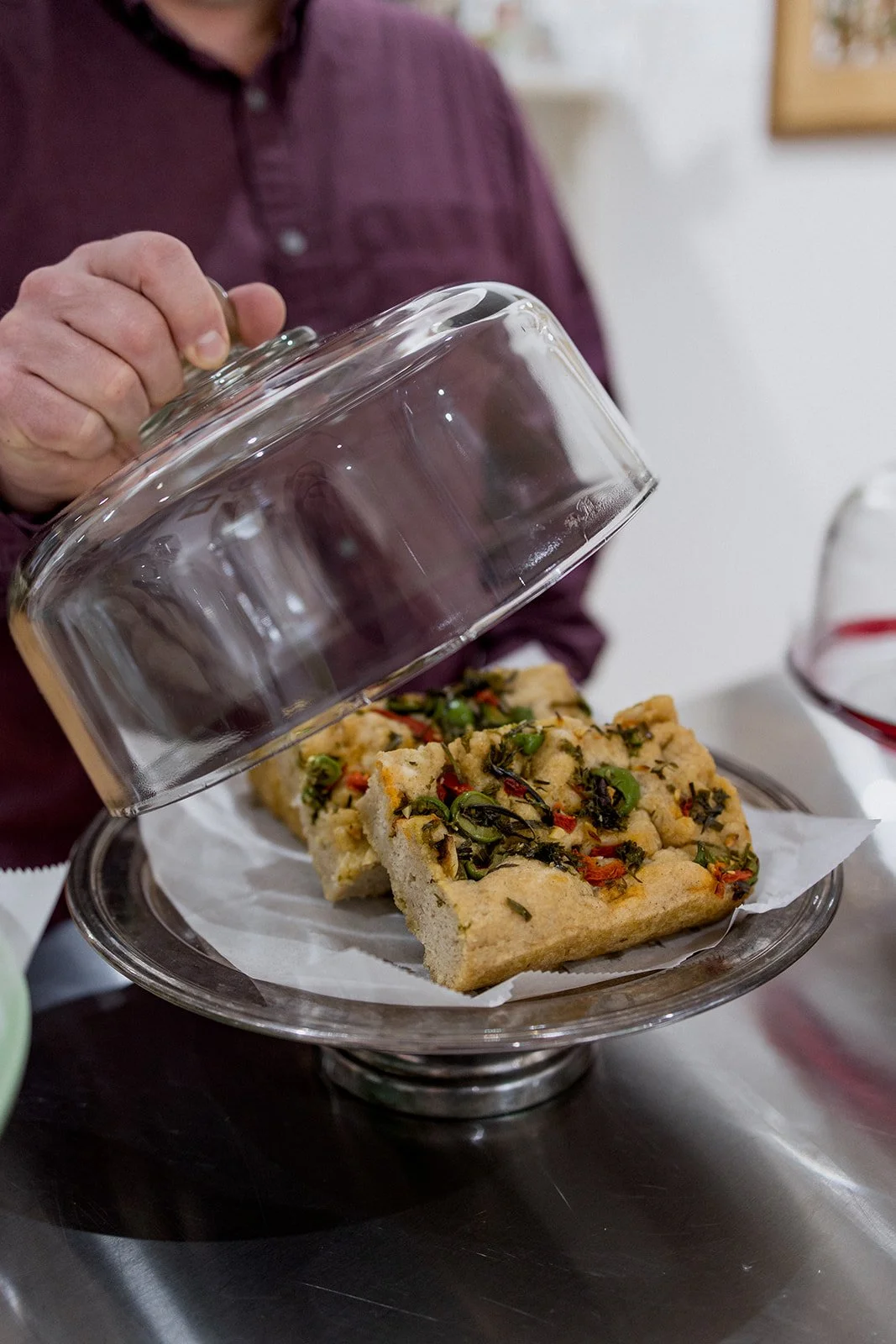 A person in a maroon shirt lifting the glass lid off a metal tray holding a rectangular focaccia bread topped with herbs, red peppers, and greens, placed on white parchment paper.