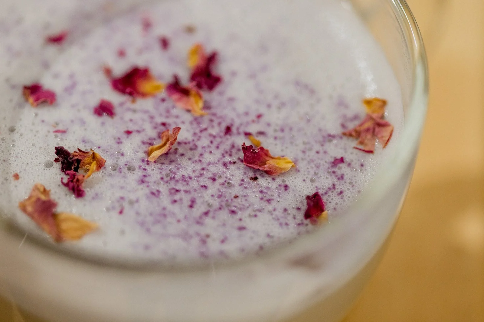 A frothy beverage garnished with dried rose petals and purple flower powder in a clear glass.