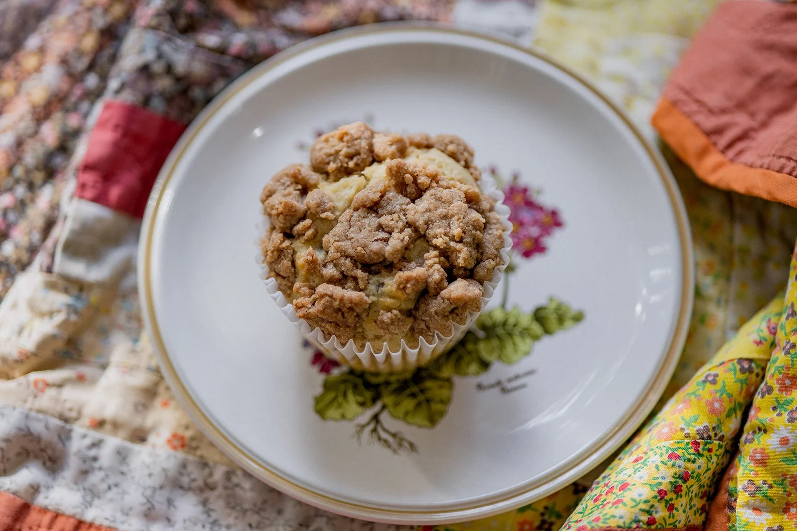 Close-up of a muffin with crumbly streusel topping on a decorative plate with floral design, surrounded by patchwork quilt fabric.