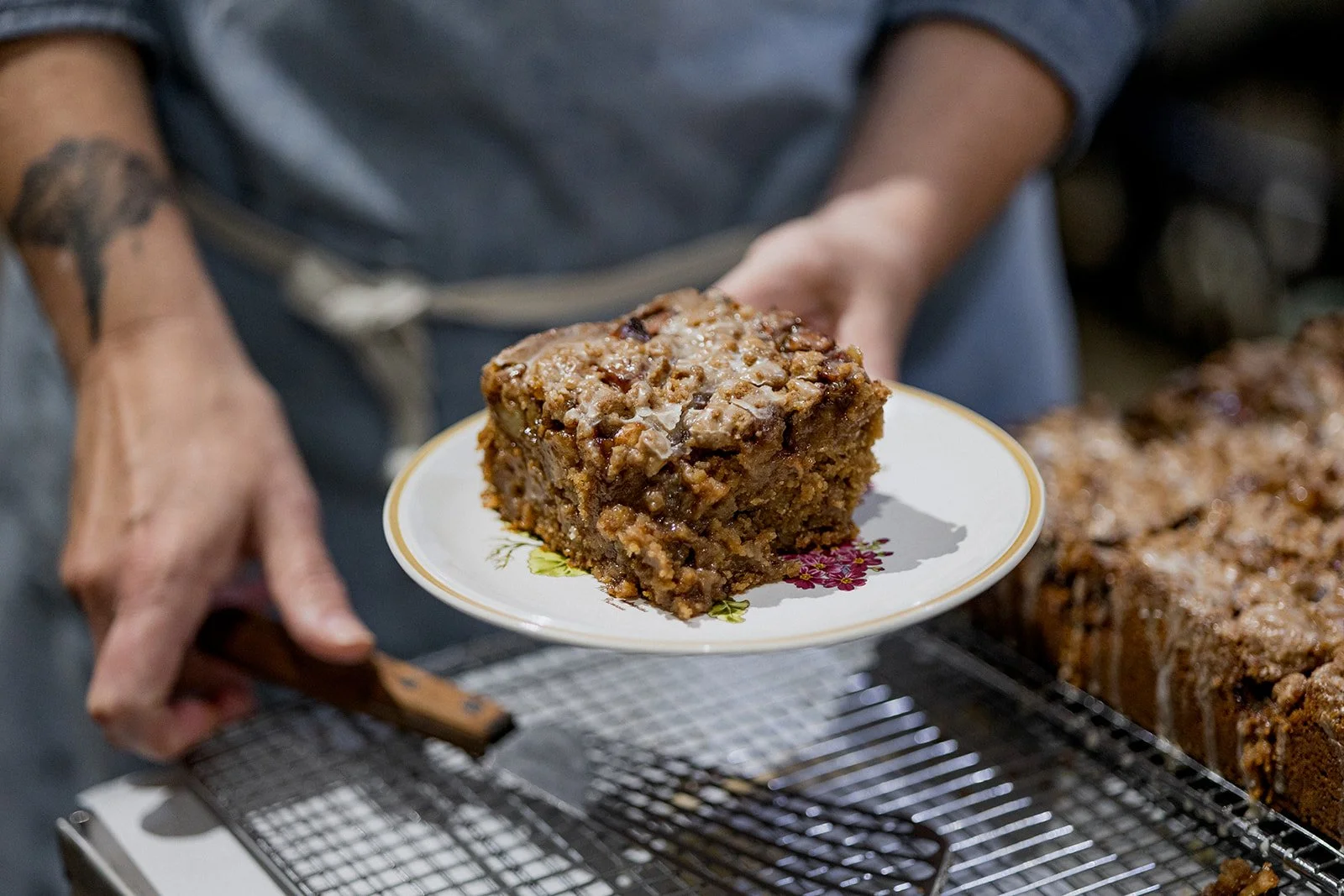 Person holding a plate with a square slice of moist fruitcake with nuts, next to a pan of more fruitcake on a cooling rack.