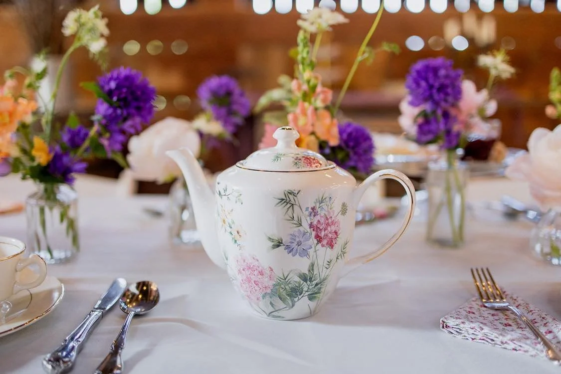 A floral teapot on a table with a white tablecloth, set for a meal with plates, cutlery, and purple and white flowers in vases in the background.