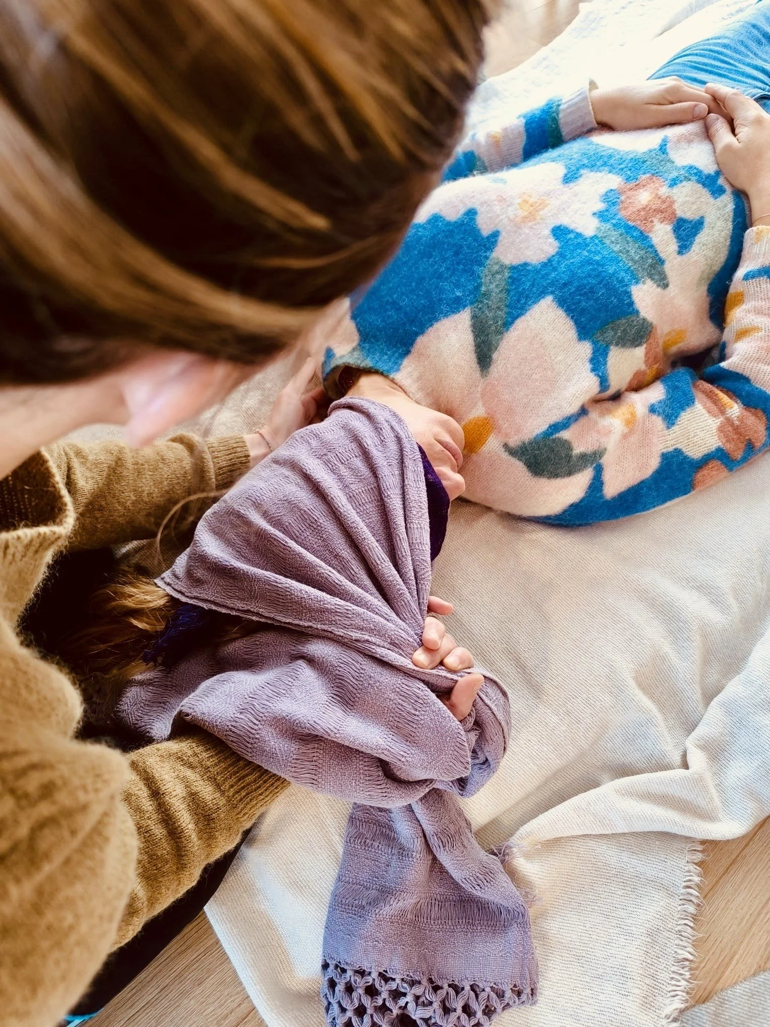 Une personne enroulée dans une couverture colorée reçoit un massage ou un soin de la personne à gauche, portant un pull marron, dans un cadre intérieur chaleureux.