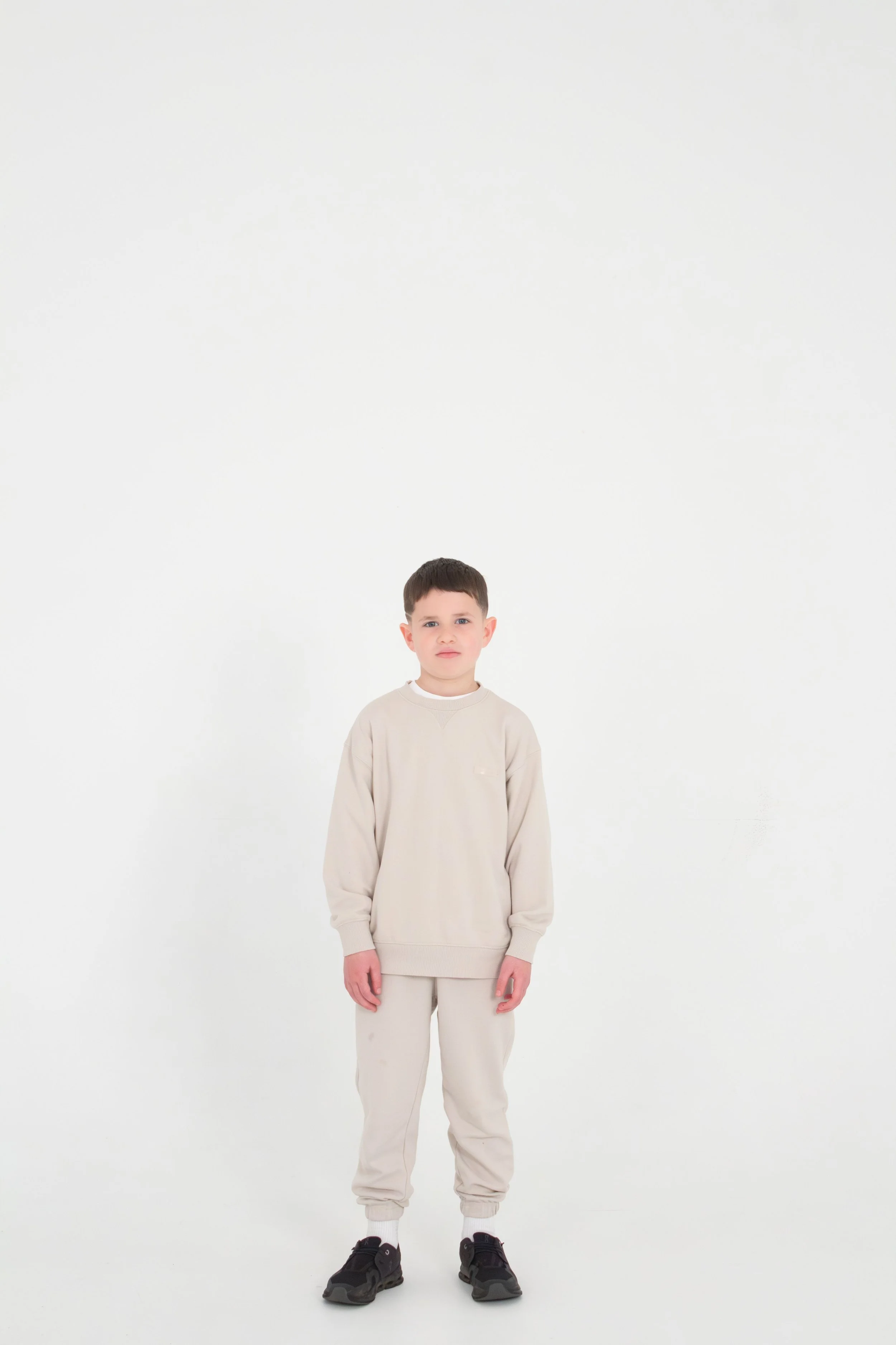 A young boy is standing against a plain white background, wearing a beige sweatshirt, matching beige pants, white socks, and black sneakers.