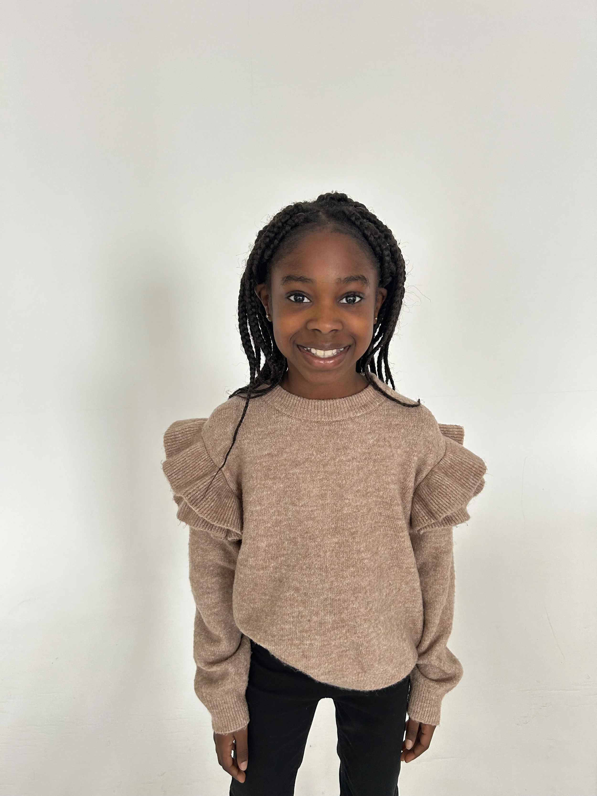 Smiling young girl with braided hair wearing a beige ruffled-sleeve sweater and black pants standing against a plain white wall. Model for leading Manchester and London based child model agency Socityuk.