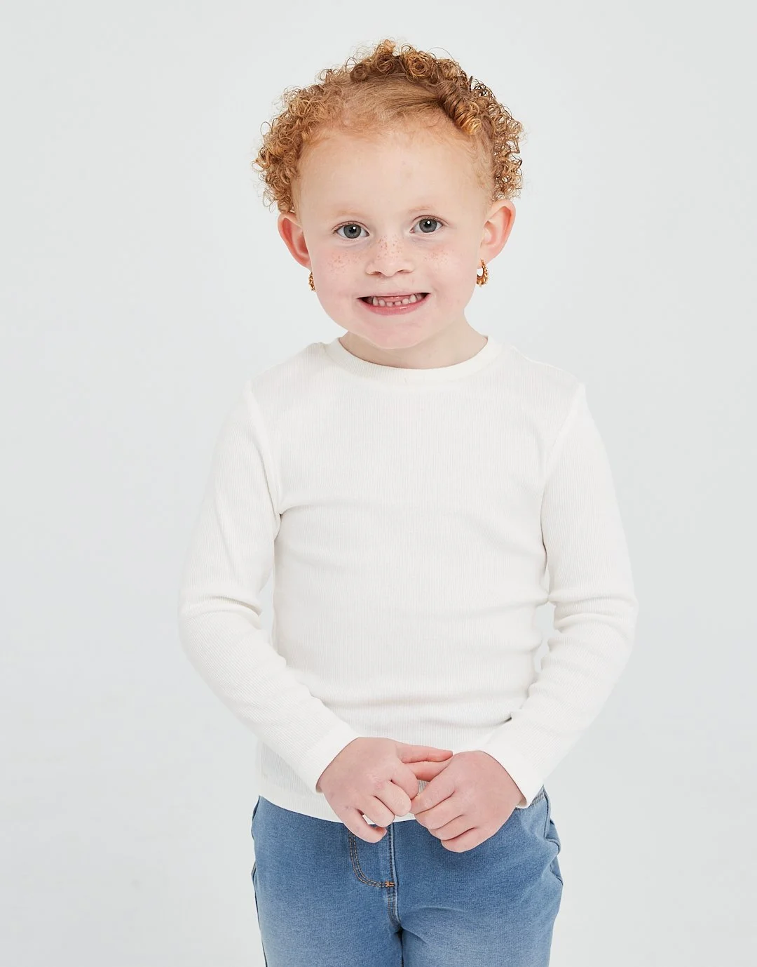A young girl with curly red hair, wearing a white long-sleeve shirt, blue jeans, and gold earrings, stands against a plain white background, smiling at the camera. Modelling for leading child model and talent agency SocityUK based in London and Manch