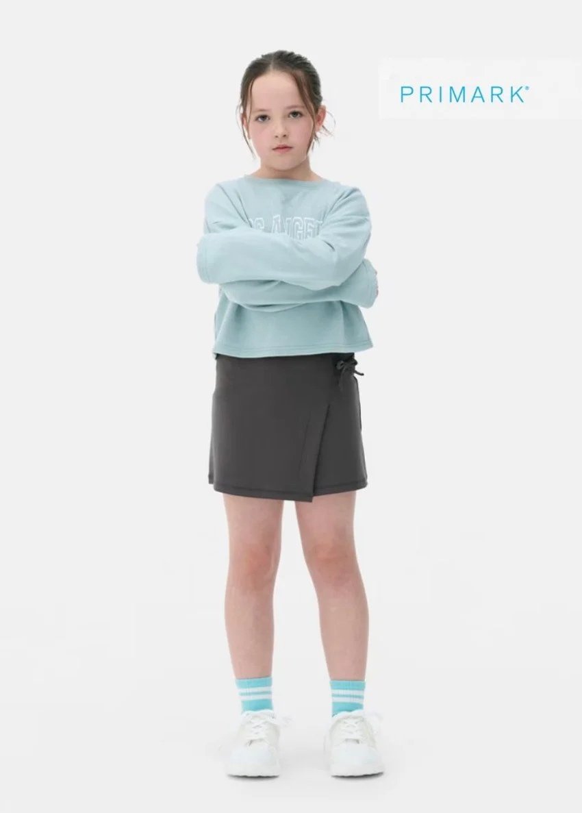 A young girl standing with arms crossed, wearing a light blue sweatshirt, a black skirt, blue and white striped socks, and white shoes, against a plain white background with the PRIMARK logo.