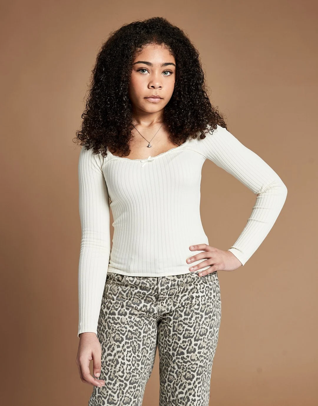 A young woman with curly dark hair, wearing a white ribbed long-sleeve top and leopard print pants, stands with one hand on her hip against a plain beige background.
