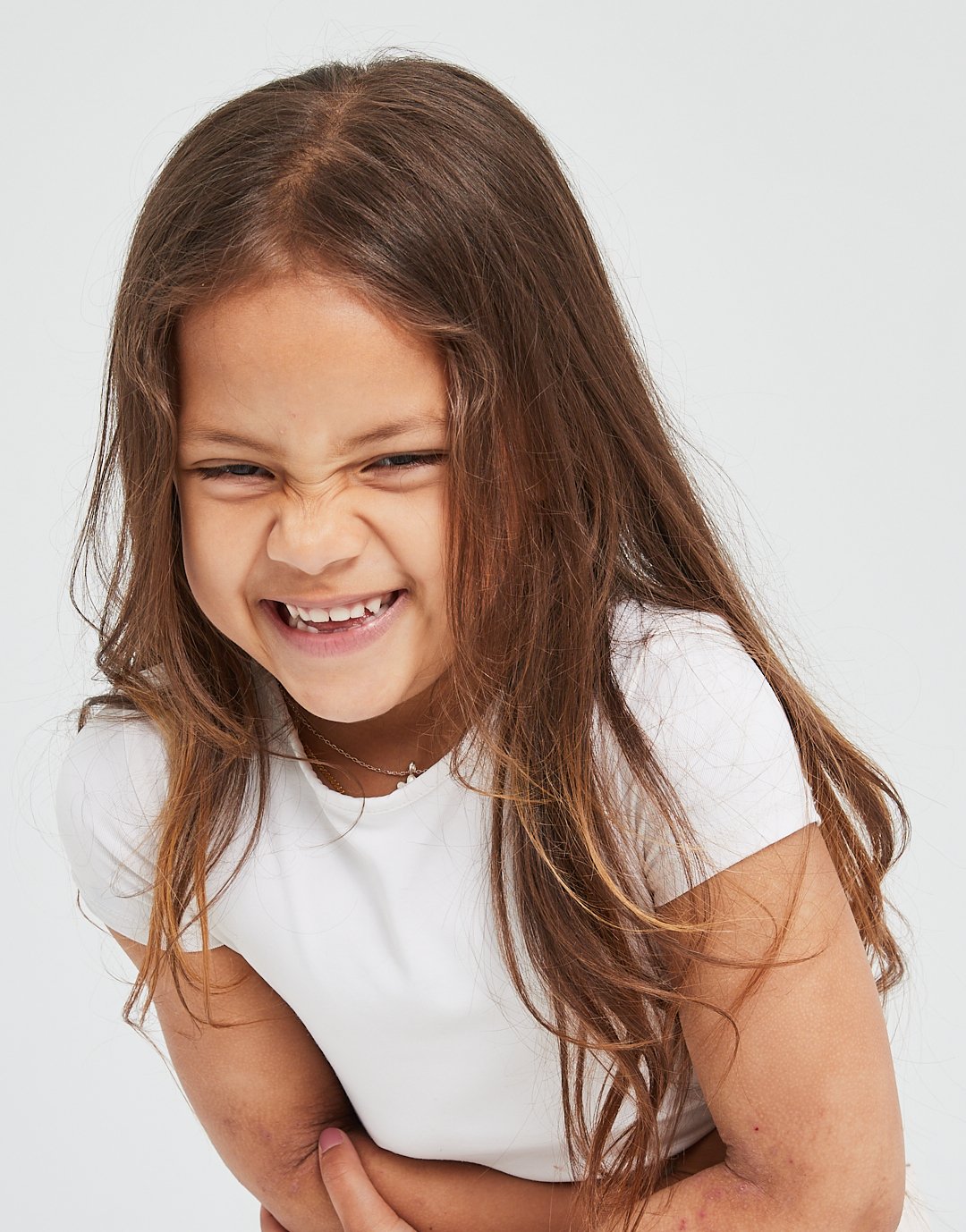 A young girl with long brown hair making a funny face, squinting her eyes and showing her teeth, wearing a white t-shirt against a plain background.