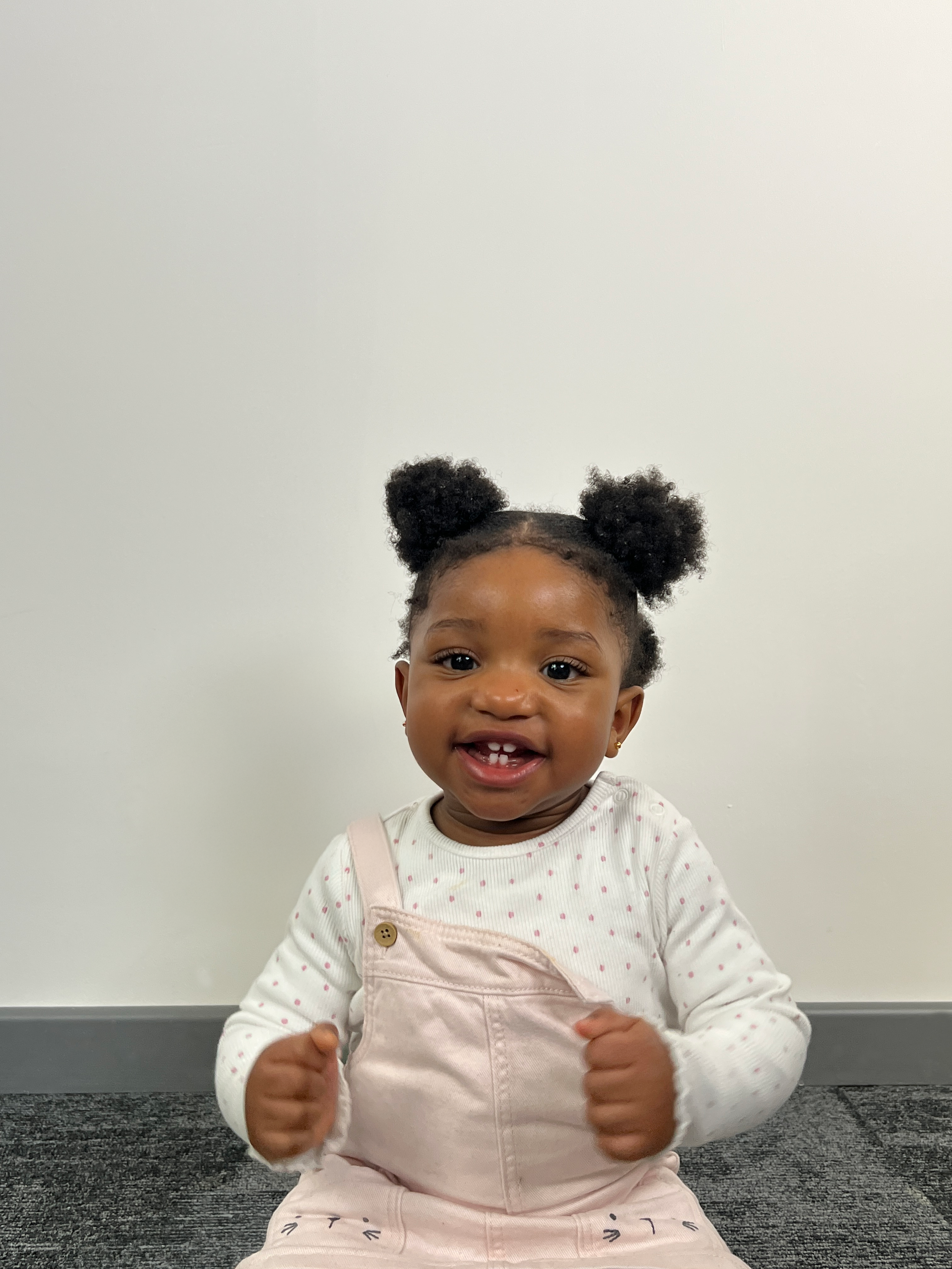 A smiling young girl with puffy hair styled in two buns, wearing a white long-sleeve shirt with small pink polka dots and pink overalls, sitting on a gray carpeted floor against a white wall.