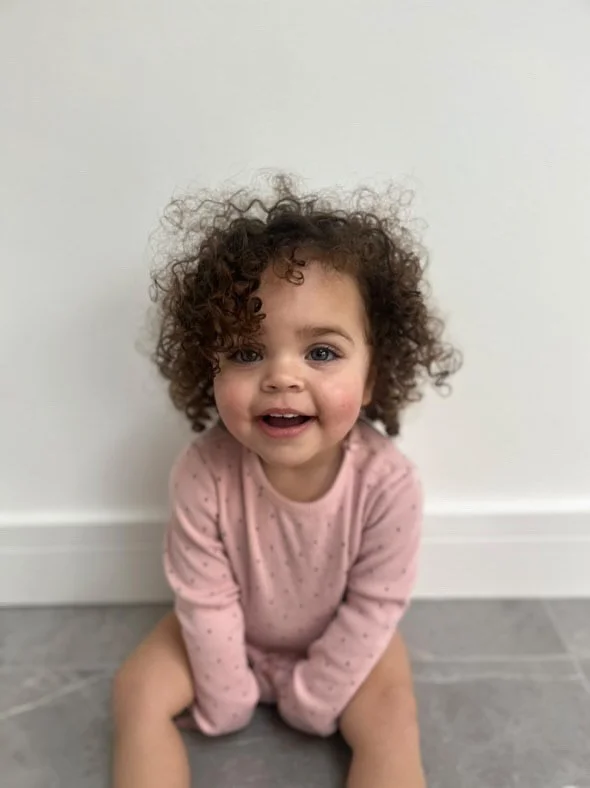A smiling toddler girl with curly hair, wearing a pink long-sleeve shirt, sitting on a gray tiled floor against a white wall. Modelling for leading child model and talent agency SocityUK based in London and Manchester.