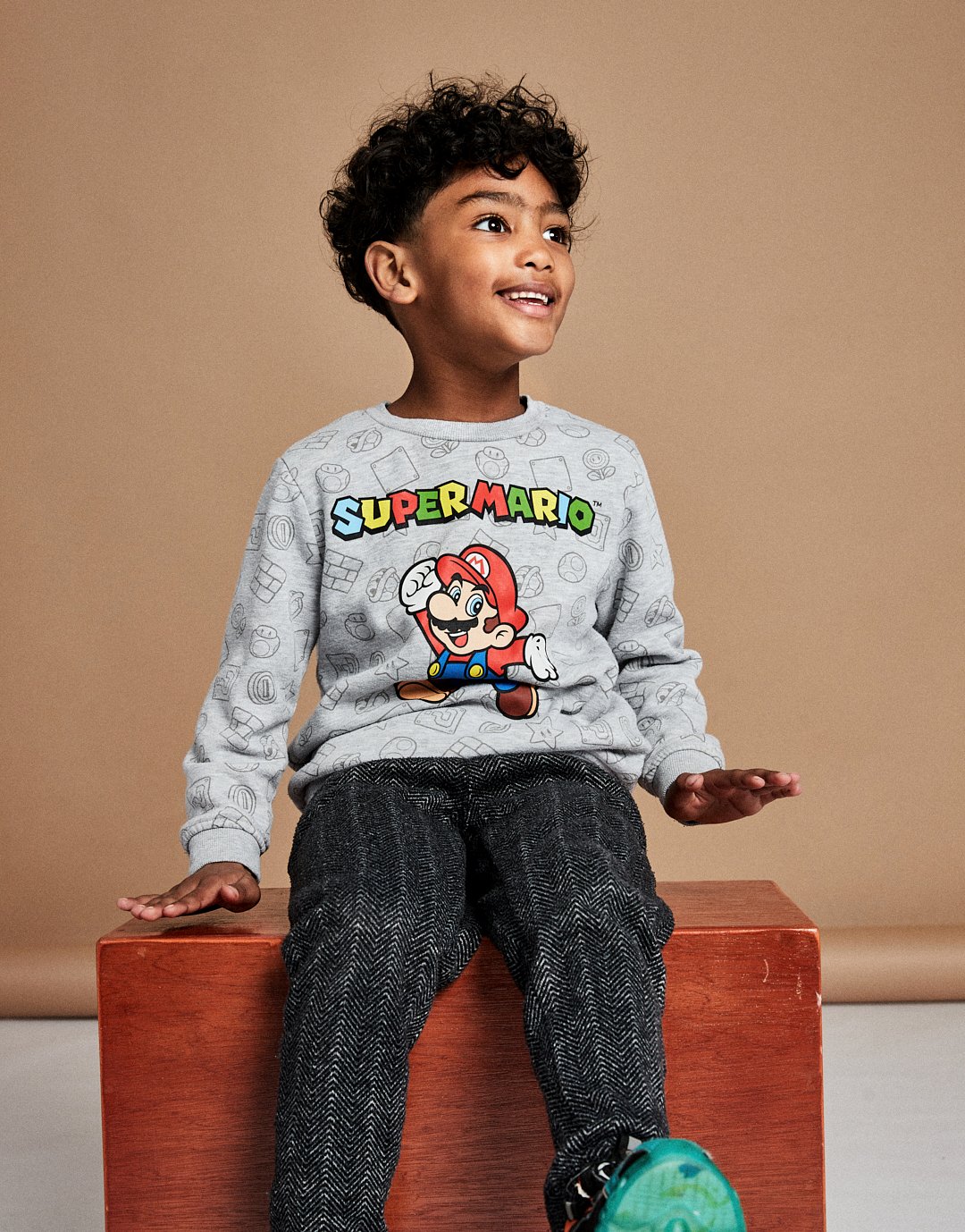 A young boy with curly black hair and brown skin is sitting on a wooden box. He is wearing a gray Super Mario sweatshirt, gray patterned trousers, and teal sneakers. He is smiling and looking to the side against a beige background.