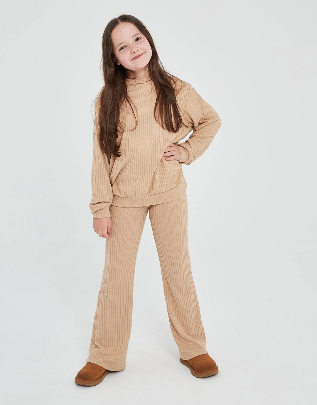 A young girl with long brown hair, wearing a beige ribbed sweatshirt and matching pants, standing on a plain light background with a confident smile, one hand on her hip.