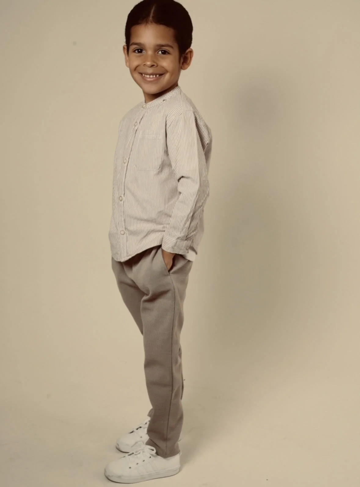 A young boy standing against a plain background, smiling at the camera, wearing a light-colored long-sleeve shirt, beige pants, and white sneakers.