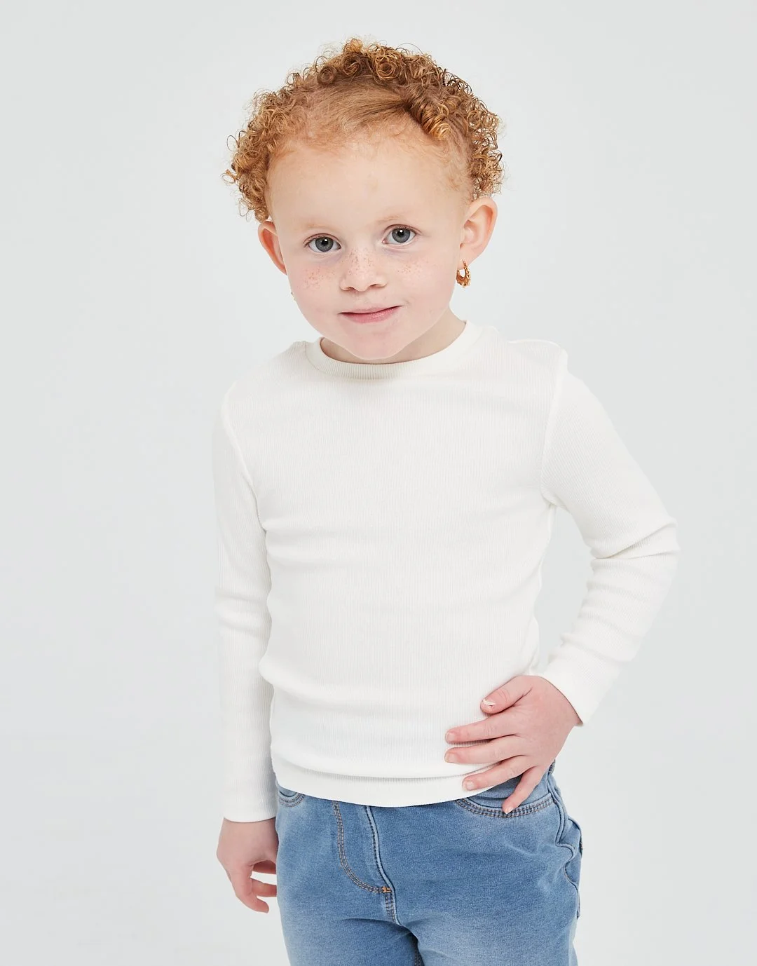 A young girl with curly red hair, blue eyes, and freckles wearing a white long-sleeve shirt and blue jeans, standing against a plain white background.