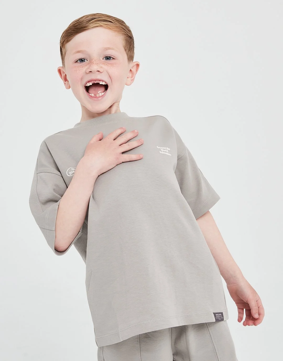 A smiling boy with red hair and freckles, wearing a gray t-shirt and shorts, standing against a plain white background.