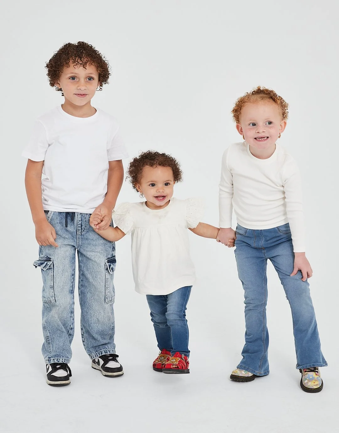 Four children holding hands and smiling, standing against a white background. They are wearing casual clothing, including jeans and white tops.Modelling for leading child model and talent agency SocityUK based in London and Manchester.