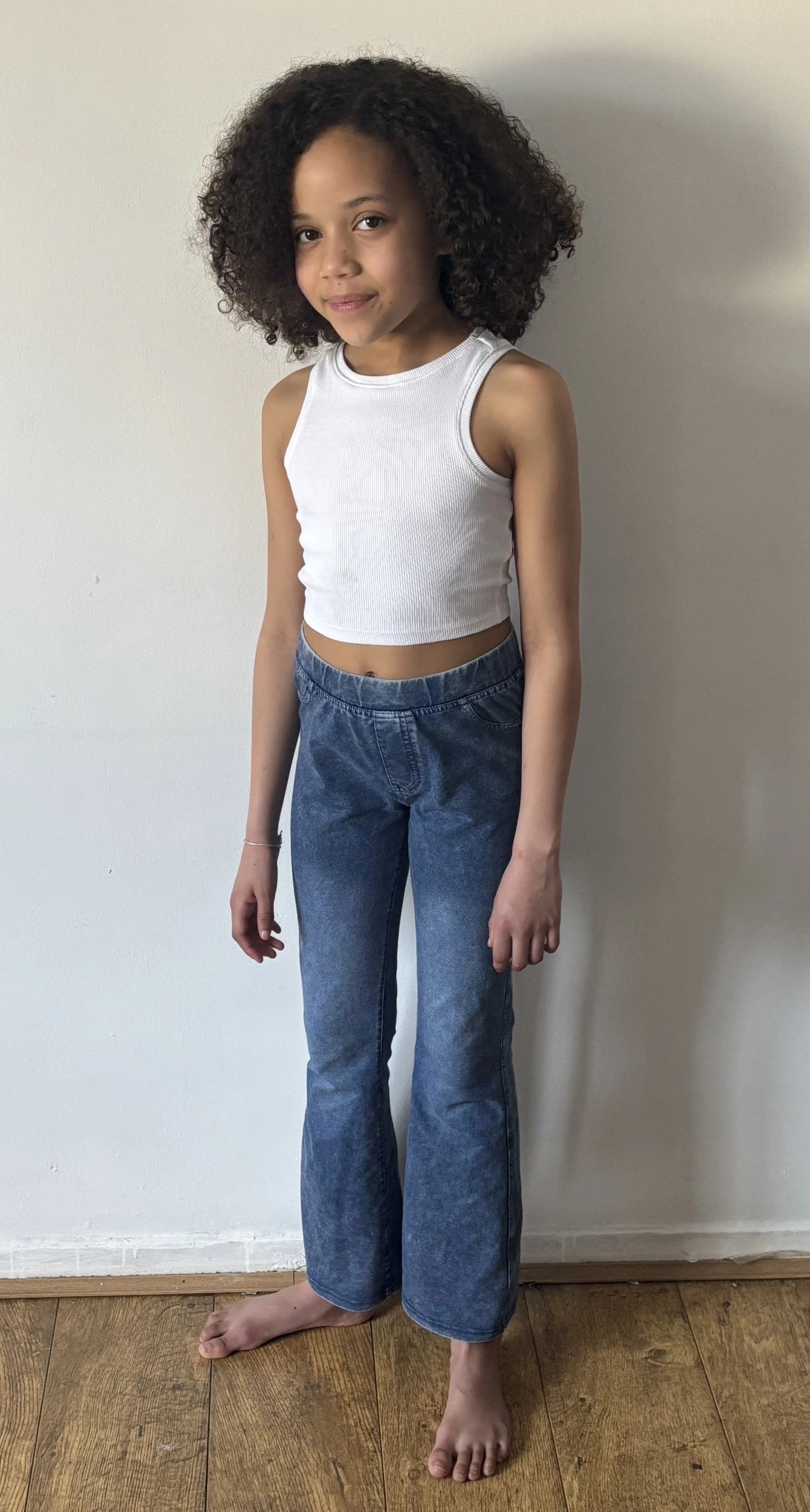 A young girl with curly hair wearing a white sleeveless crop top and blue jeans standing barefoot indoors against a plain wall.