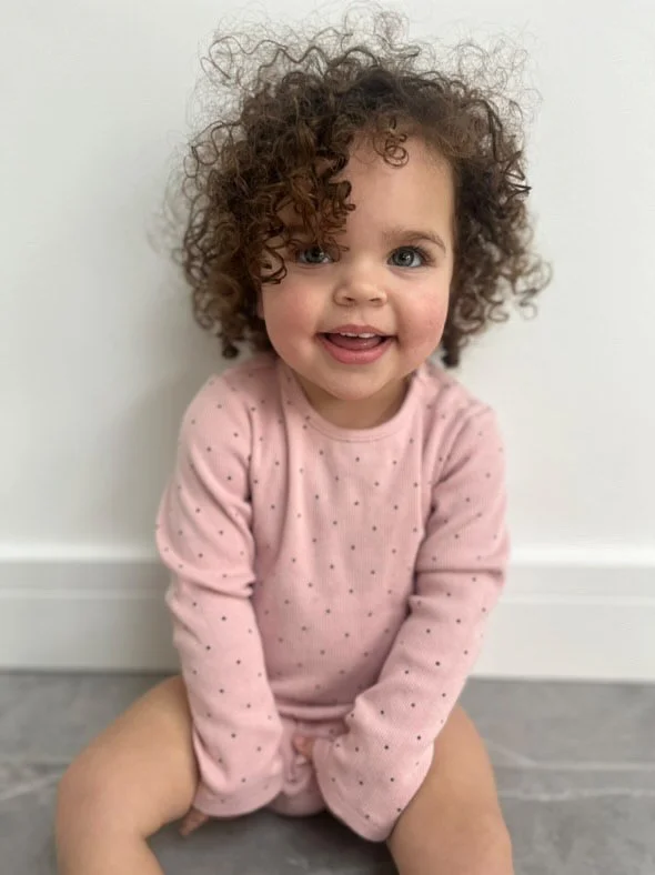 A young girl with curly hair and blue eyes, wearing a pink long-sleeve shirt with small dots, sitting on the floor against a white wall and smiling.Modelling for leading child model and talent agency SocityUK based in London and Manchester.
