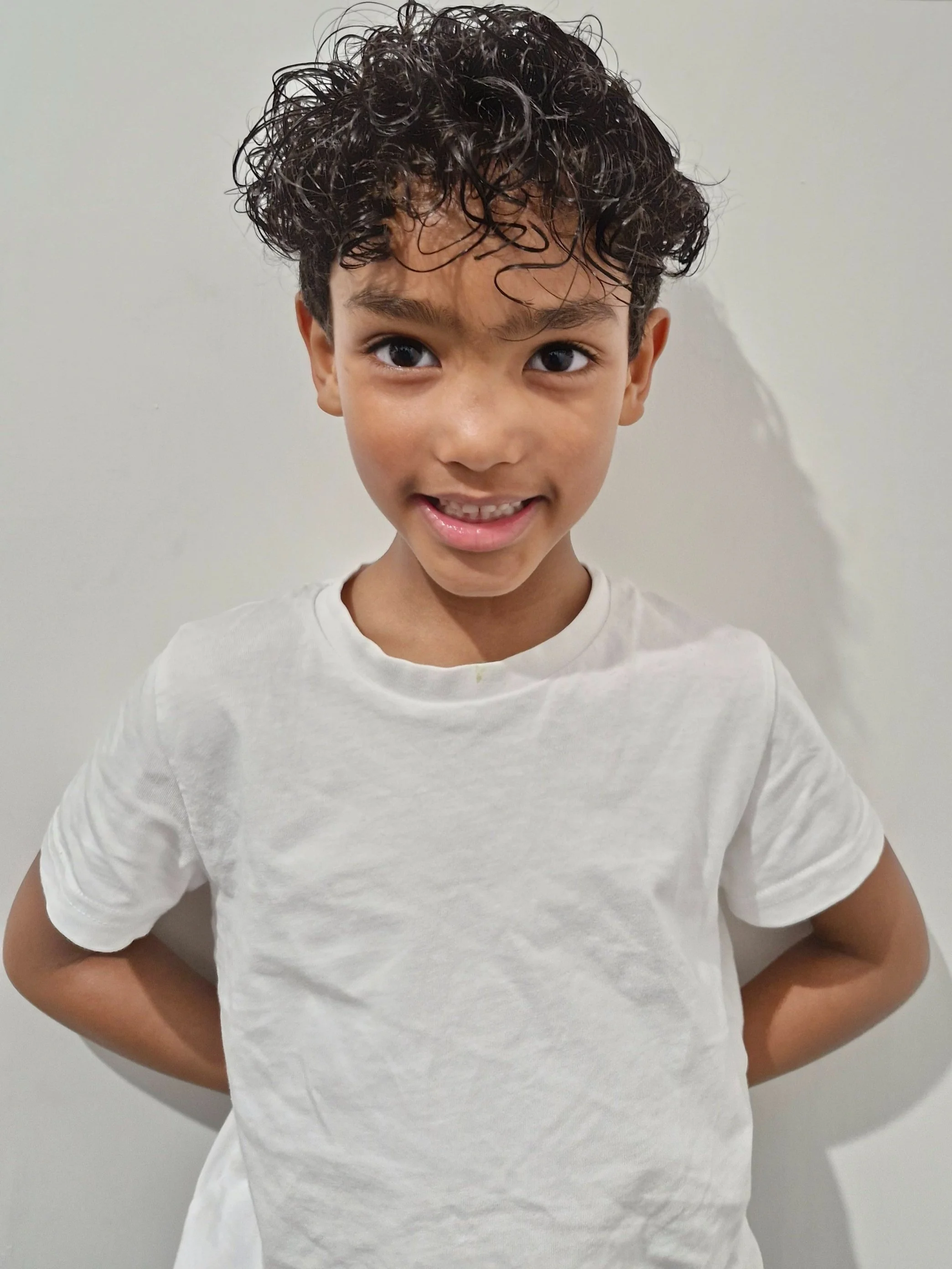 A smiling young boy with dark curly hair, wearing a plain white t-shirt, standing against a light-colored wall. Modelling for leading child model and talent agency SocityUK based in London and Manchester.