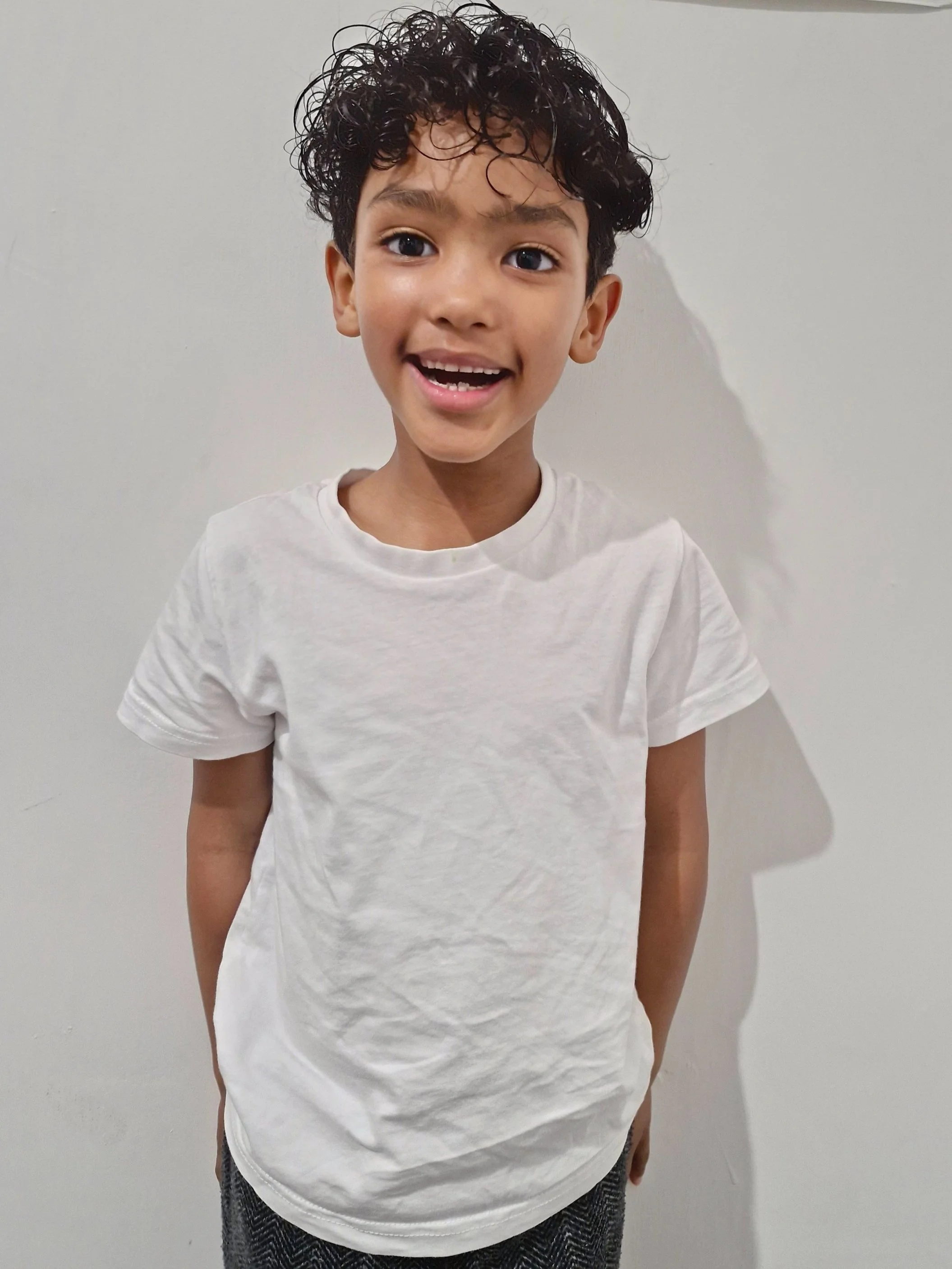 A young boy with curly dark hair smiling, wearing a white T-shirt, standing against a plain light-colored wall. Modelling for leading child model and talent agency SocityUK based in London and Manchester.