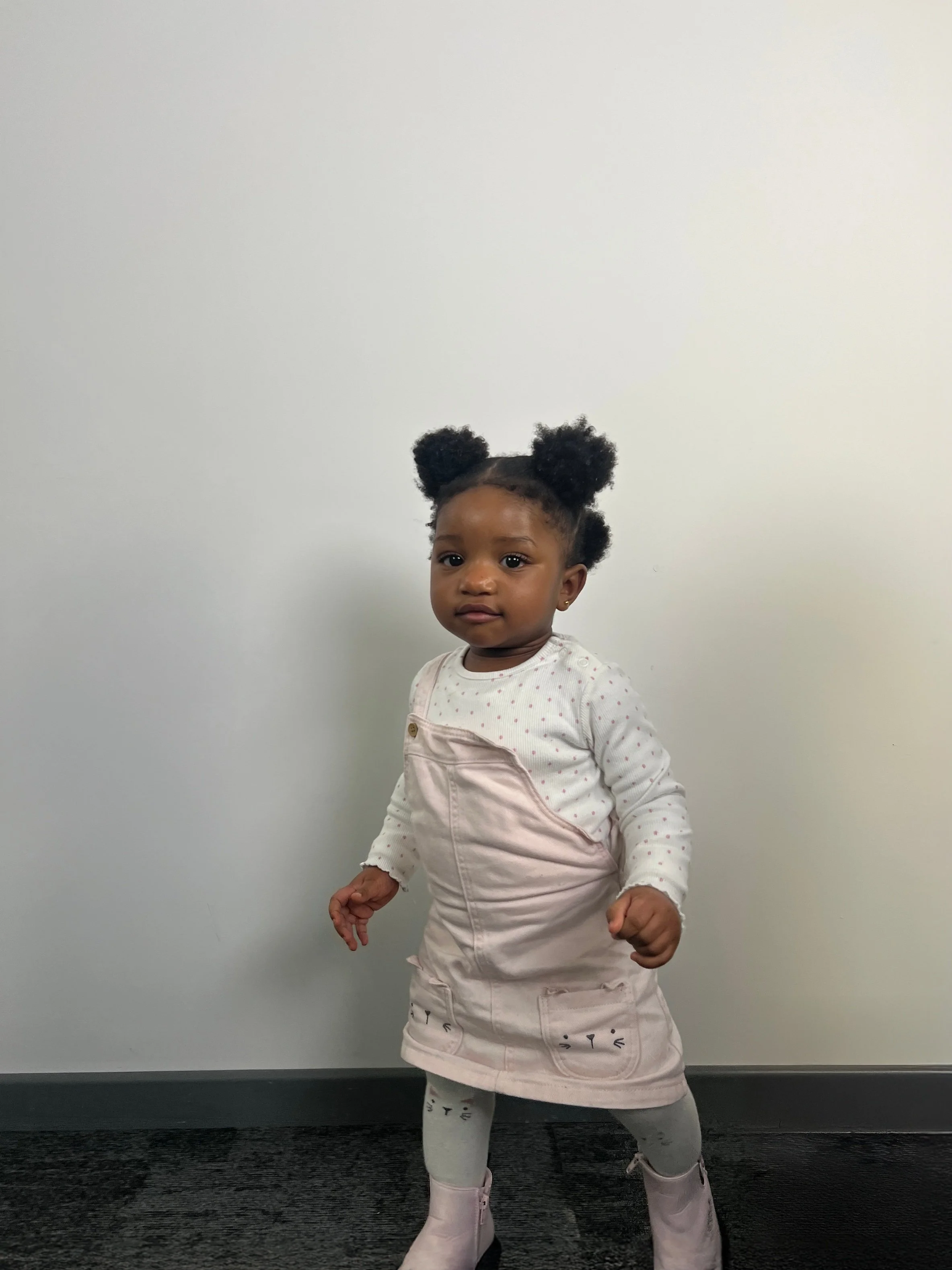 A young girl with dark curly hair in two puffs, wearing a pink pinafore dress layered over a white polka dot long-sleeve shirt, light grey leggings with cat face designs, and light pink boots, standing against a plain off-white wall.