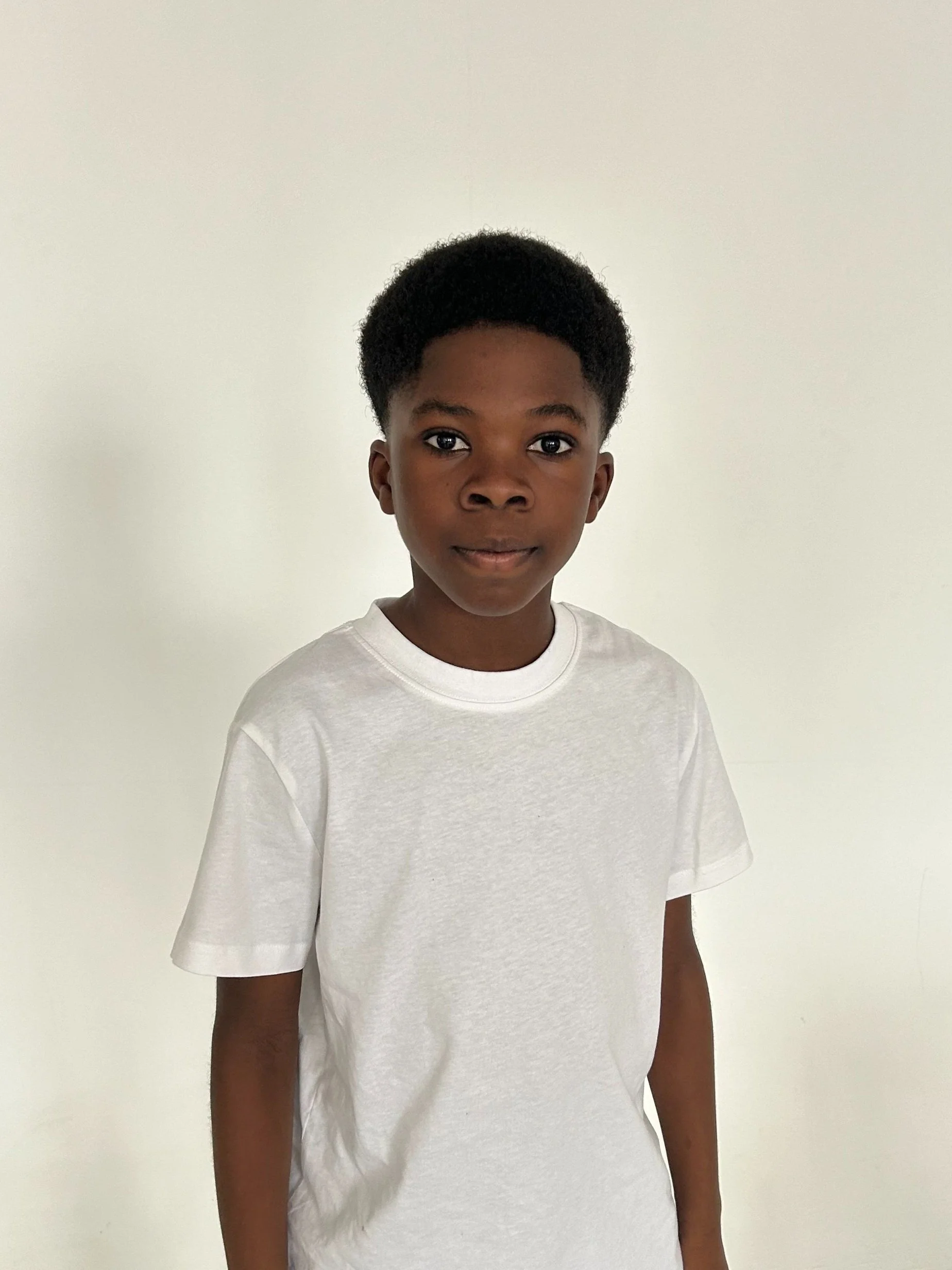A young boy with short curly hair wearing a plain white t-shirt standing in front of a plain white wall.