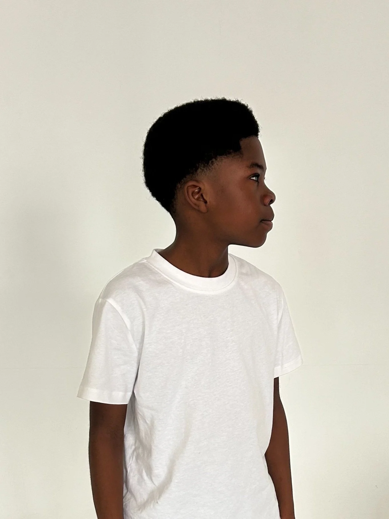A young boy with dark skin and a tall Afro hairstyle, wearing a plain white T-shirt, standing against a plain light-colored wall, looking to the right.