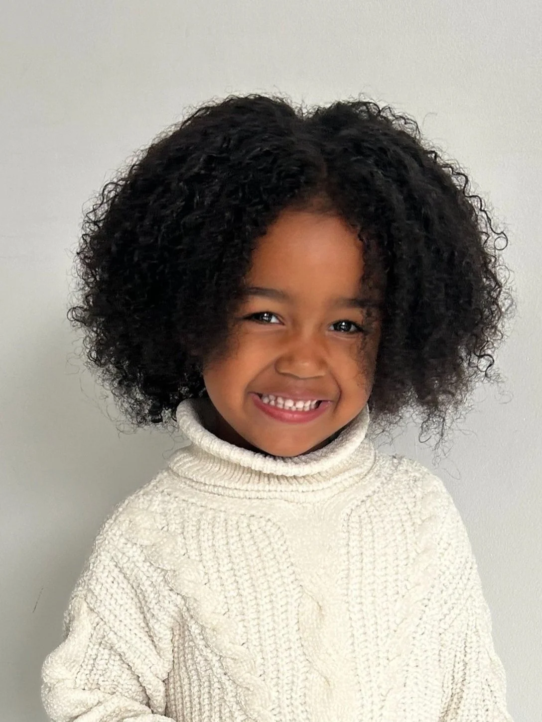 Young girl with curly black hair smiling, wearing a cream-colored turtleneck sweater, standing against a light-colored wall. Model for leading Manchester and London based child model agency Socityuk.