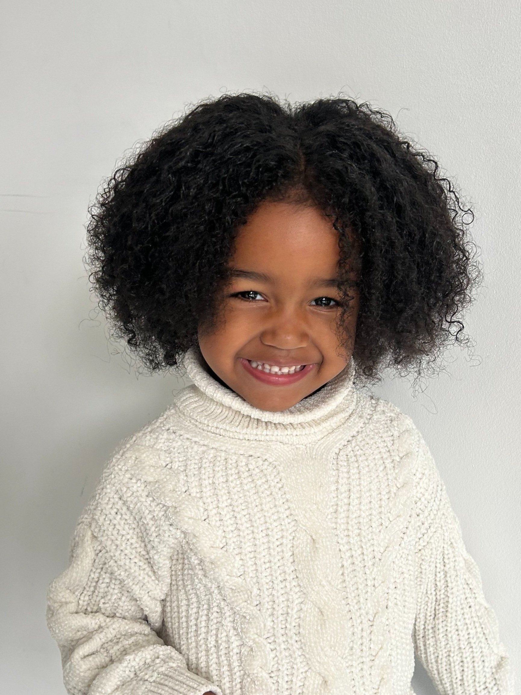 Close-up of a young girl with dark curly hair smiling, wearing a cream-colored knit turtleneck sweater, standing in front of a plain light-colored wall. Model for leading Manchester and London based child model agency Socityuk.