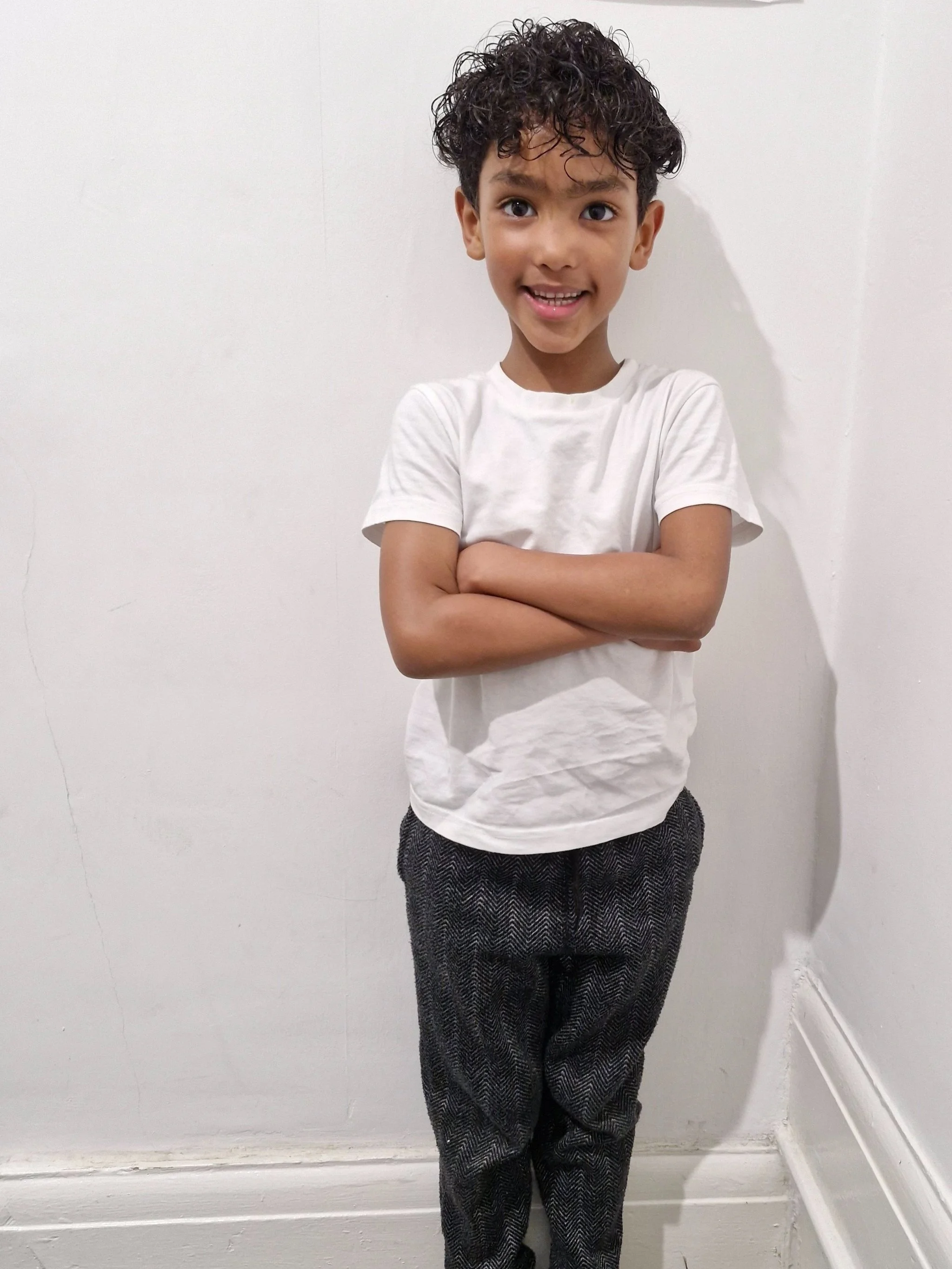 A young boy with curly dark hair, smiling and crossing his arms, standing against a plain light-colored wall. Modelling for leading child model and talent agency SocityUK based in London and Manchester.