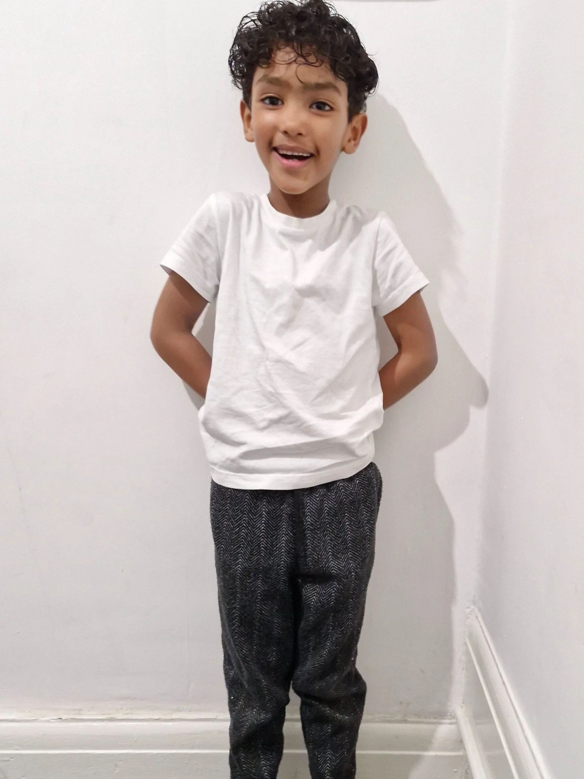 A young boy with curly hair smiling and standing against a plain white wall, wearing a white t-shirt and patterned dark trousers. Modelling for leading child model and talent agency SocityUK based in London and Manchester.